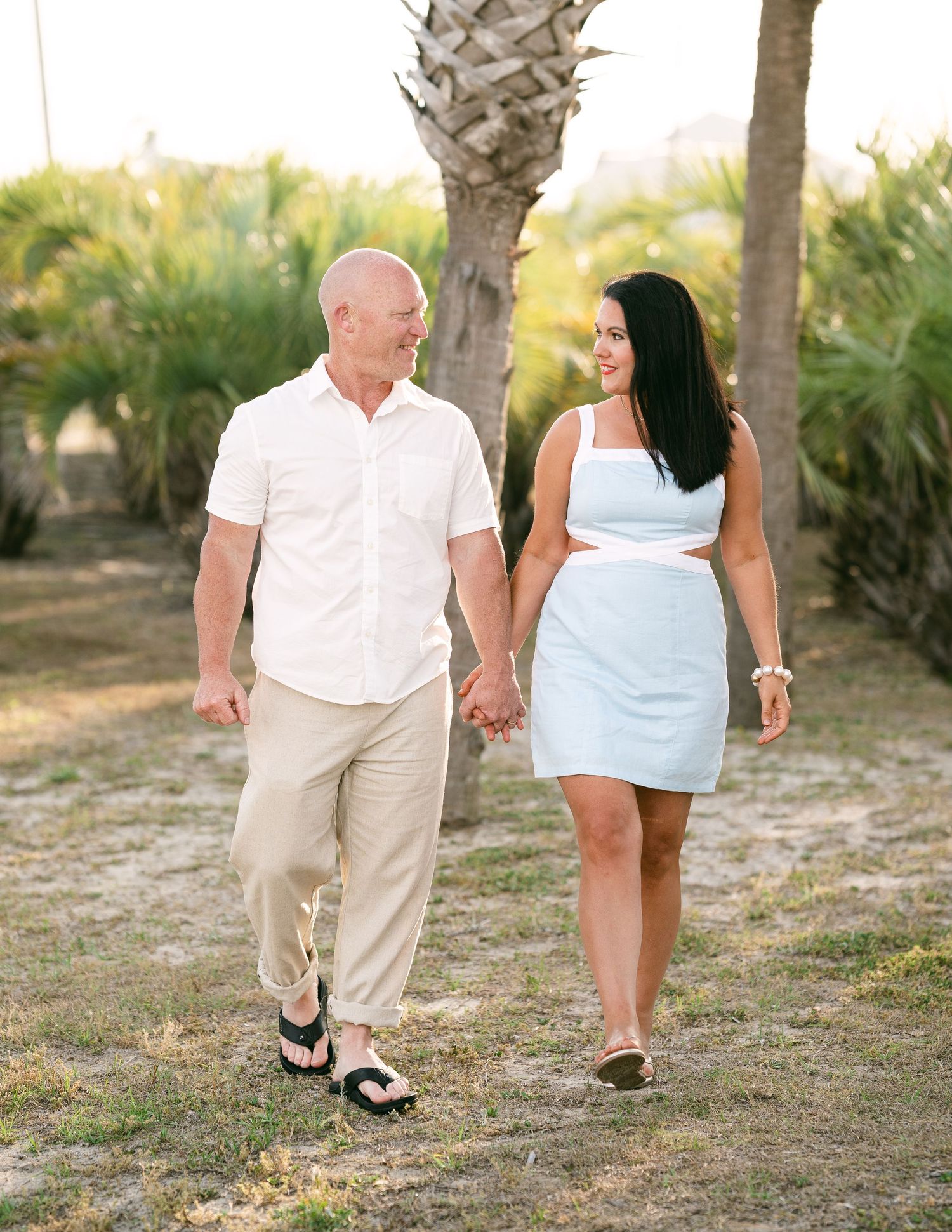 Couple holding hands walking through palm-lined coastal path in coordinated white and light blue outfits.