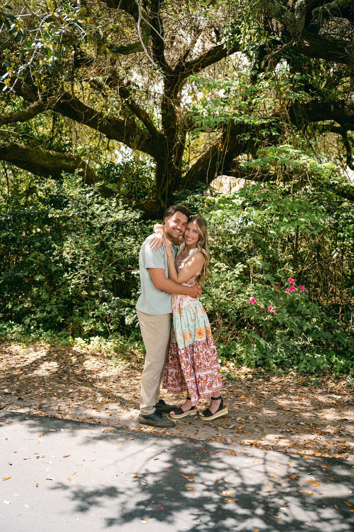 Couple hugs warmly beneath a large mossy oak tree in a lush garden.