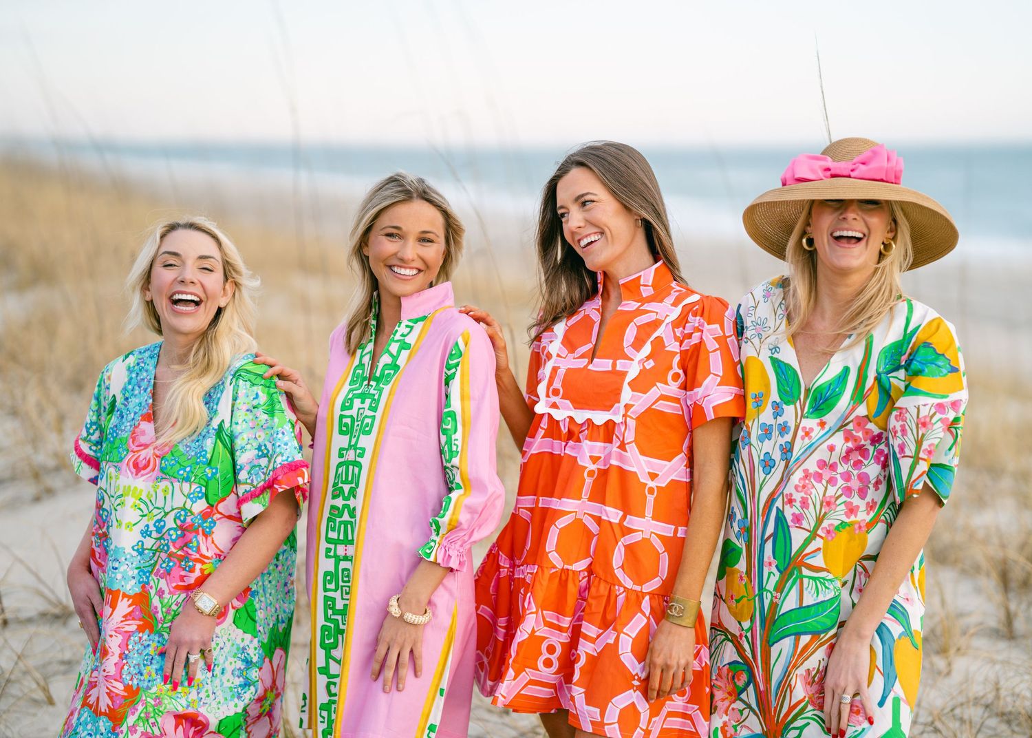 Four women laughing in colorful printed dresses on a sunny beach.