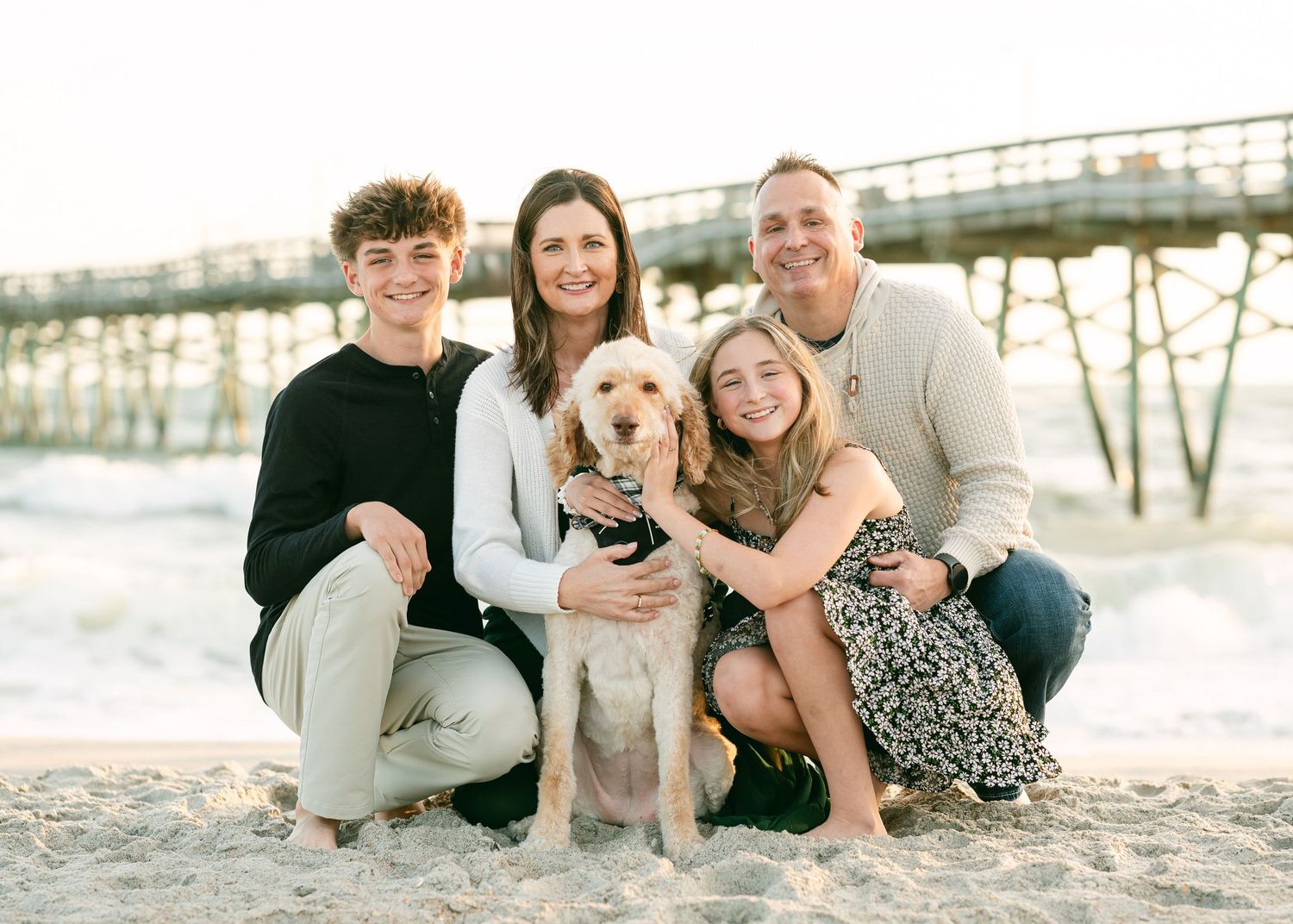 Family of four with golden dog posing on beach near pier at sunset.