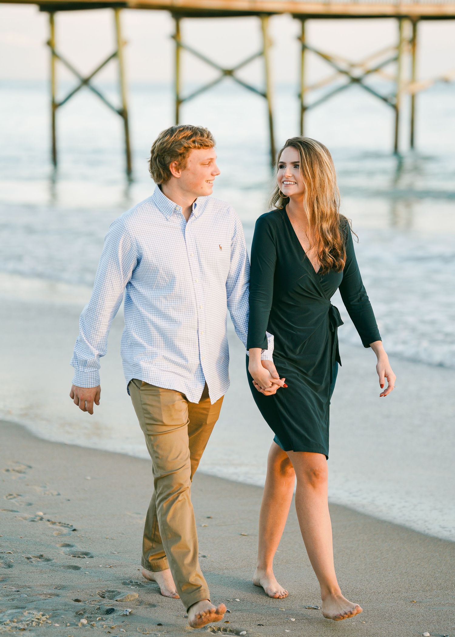 Couple walking hand in hand barefoot on the beach near a pier at golden hour.