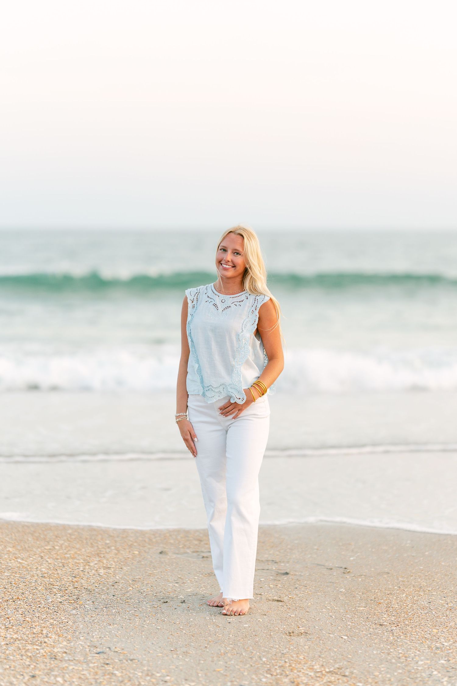 Smiling blonde woman in light blue top and white pants standing barefoot on a sandy beach.
