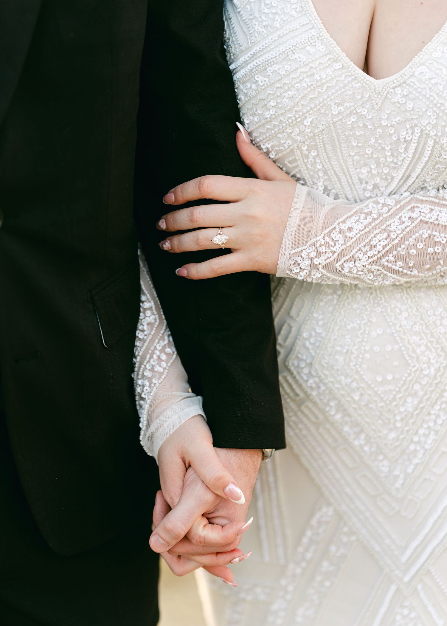 Close-up of bride in beaded lace gown holding grooms hand showing stunning emerald cut engagement ring.