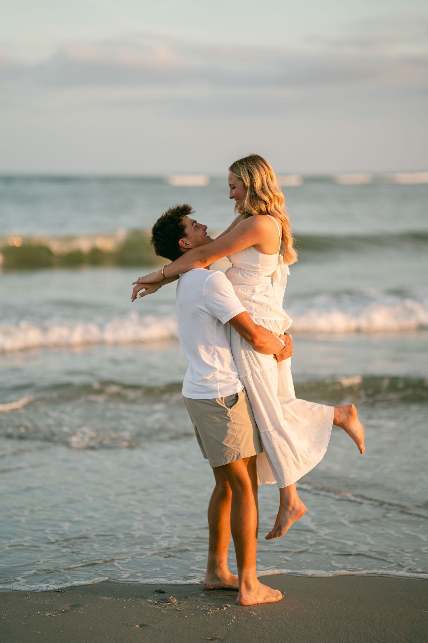 Joyful man lifting woman in white dress on sunny beach with ocean waves in background.