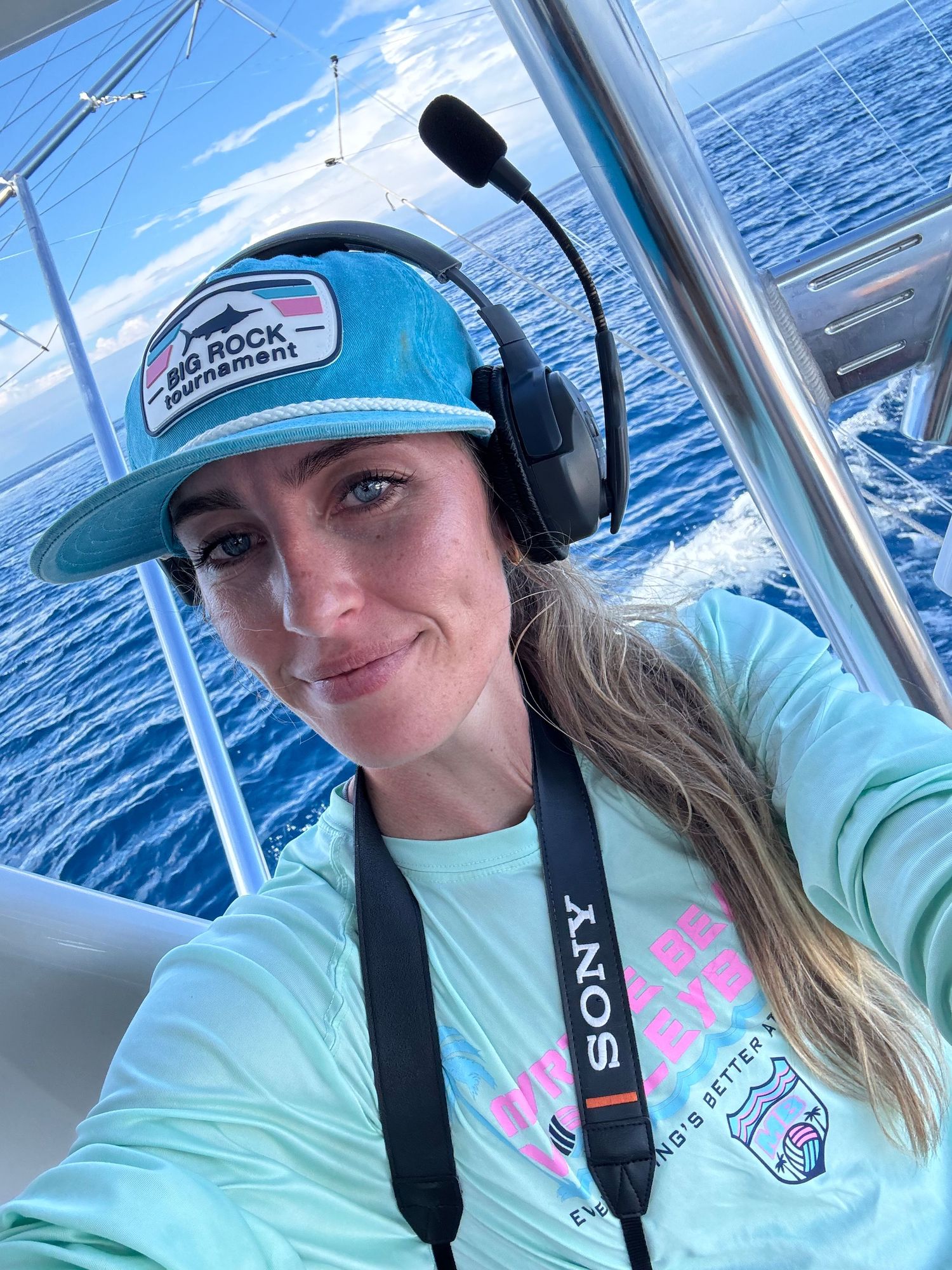 Woman wearing Sony headset and teal cap taking selfie on boat at sea.