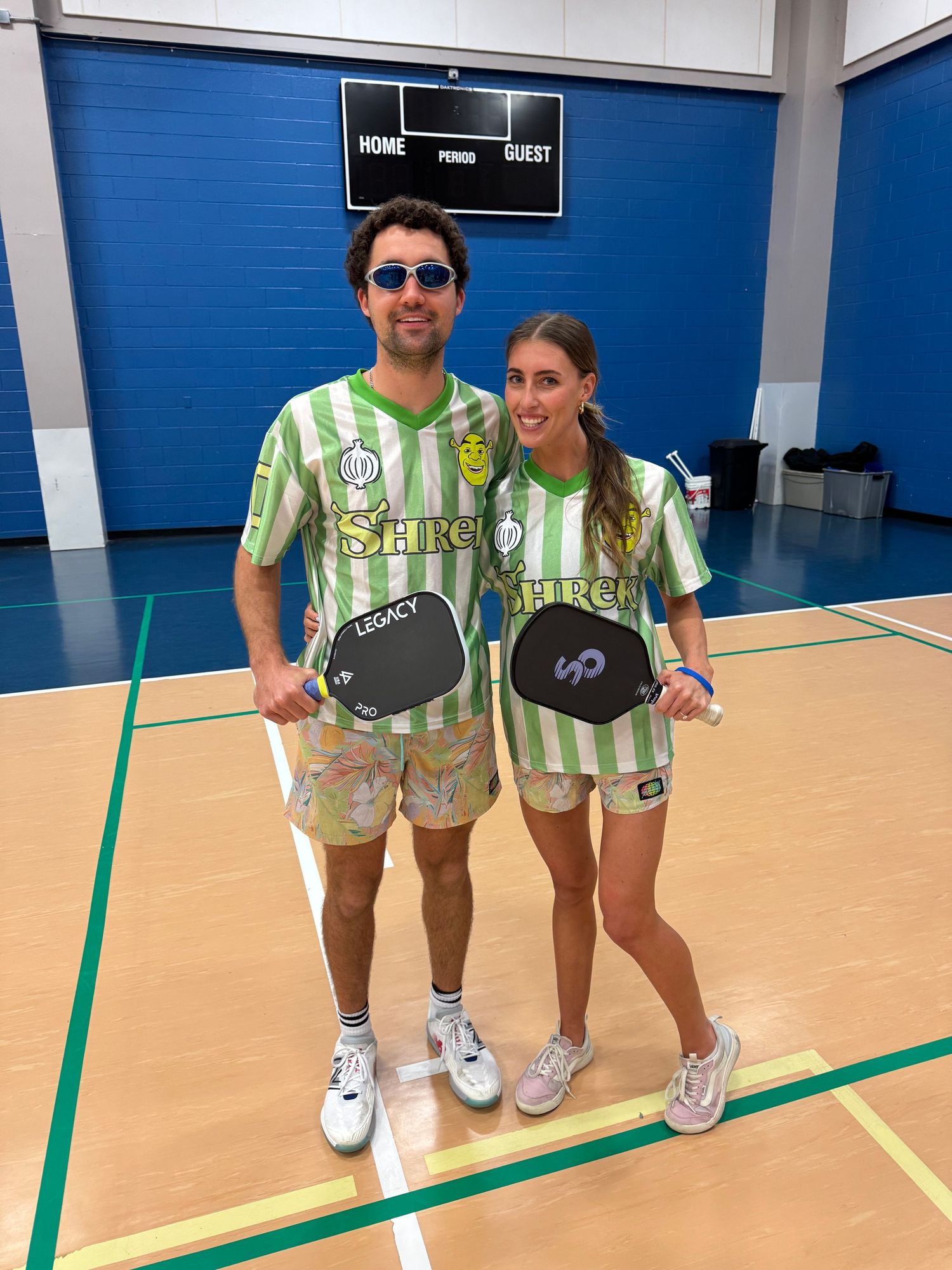 Couple in Shrek jerseys holding pickleball paddles on indoor court.