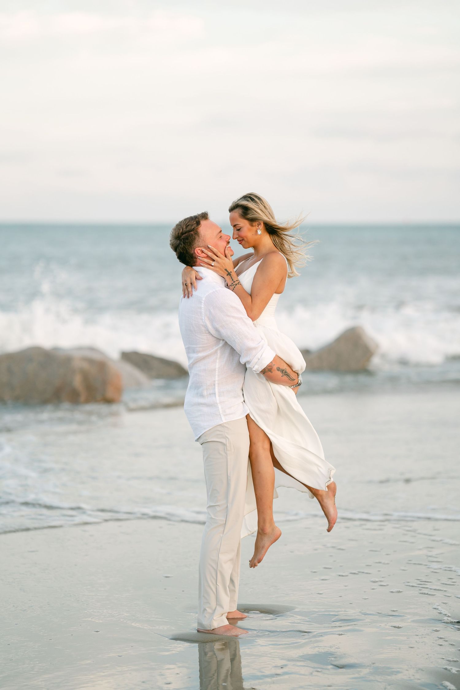 Couple in white embracing and kissing on a beach with ocean waves and rocks in the background.