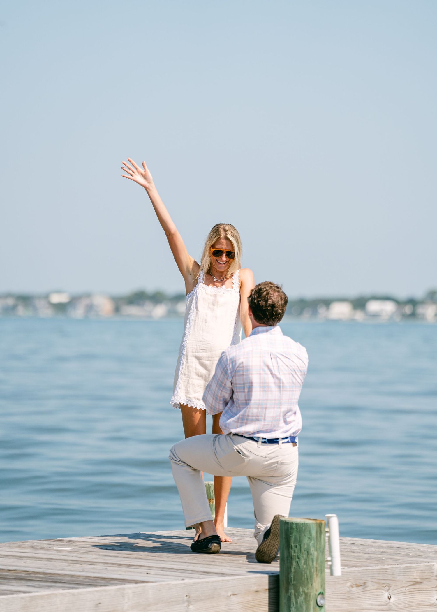 Woman raising her arm in excitement as man proposes on a wooden dock over calm water.