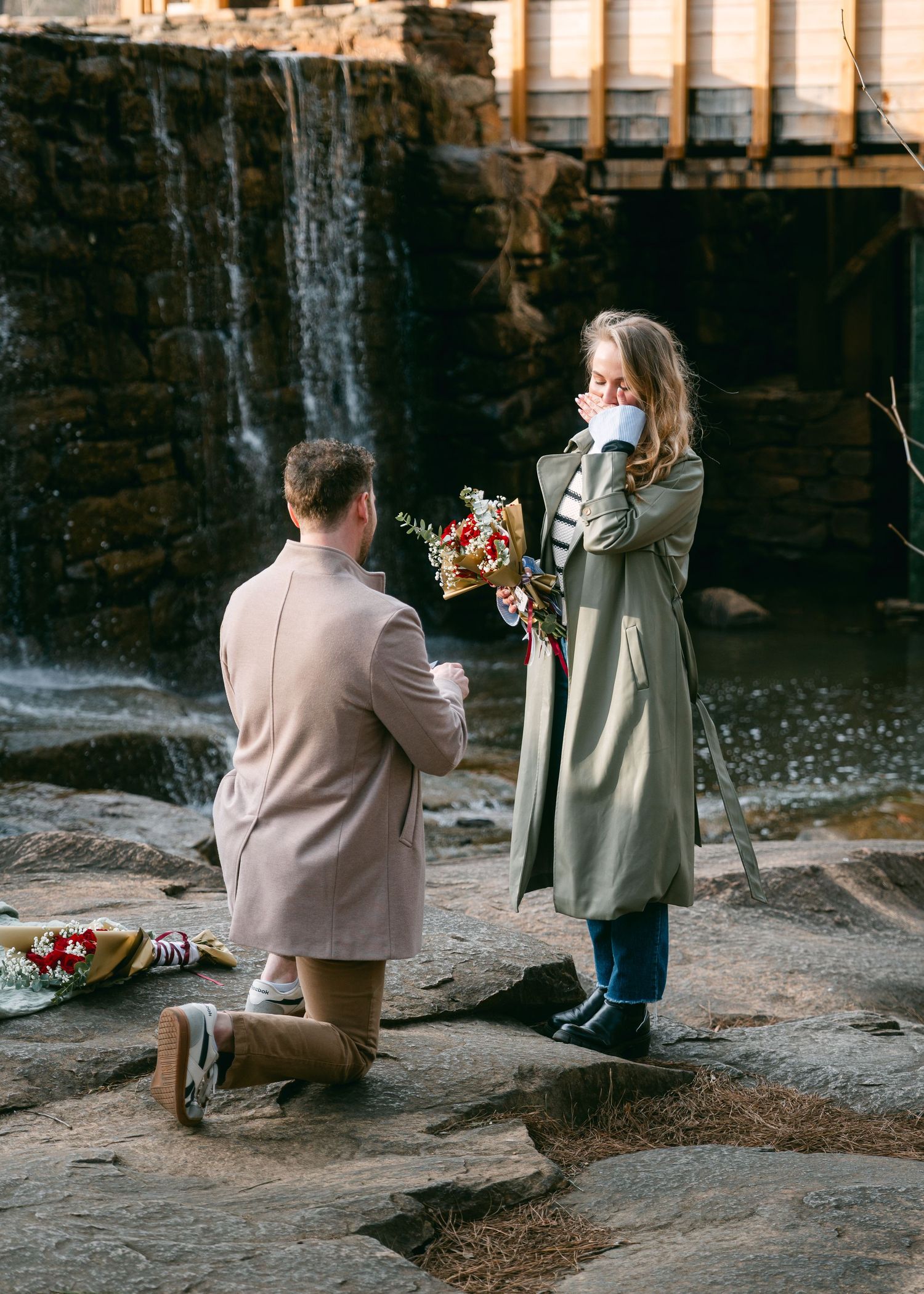 Man kneeling on rocks proposing to emotional woman with bouquet near scenic waterfall backdrop.
