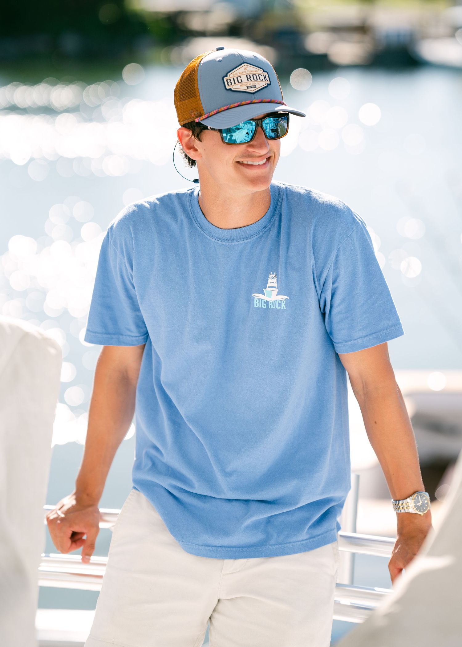 Young man wearing a blue Big Rock t-shirt and trucker hat smiles on a boat with sparkling water behind him.
