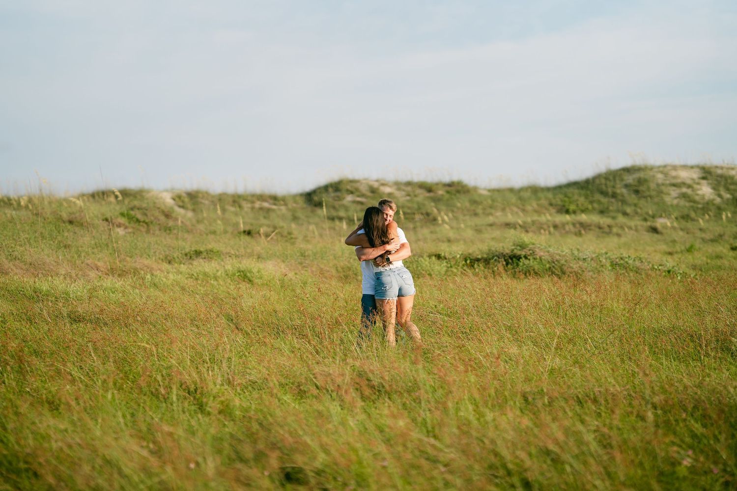 Couple embracing in a wide open green grassy field under a bright sky.