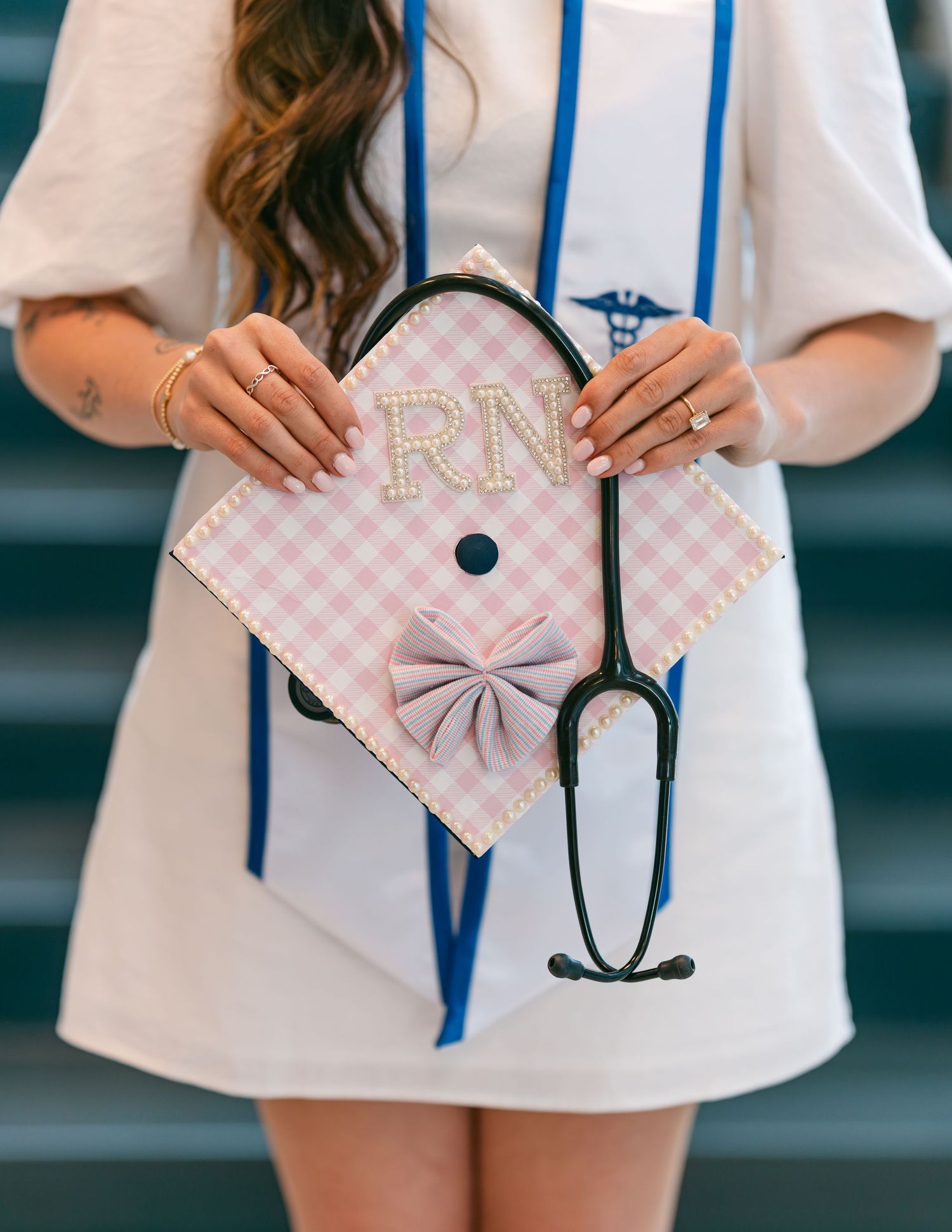 Nursing graduate holds RN decorated pink gingham graduation cap with stethoscope draped over.