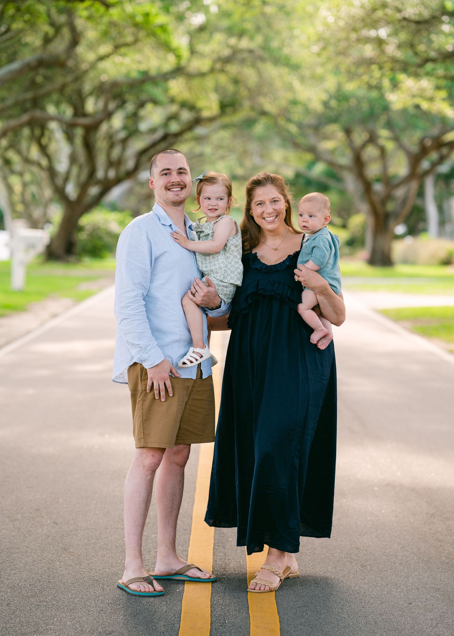 Young family of four posing on tree-lined road, parents holding toddler and baby.
