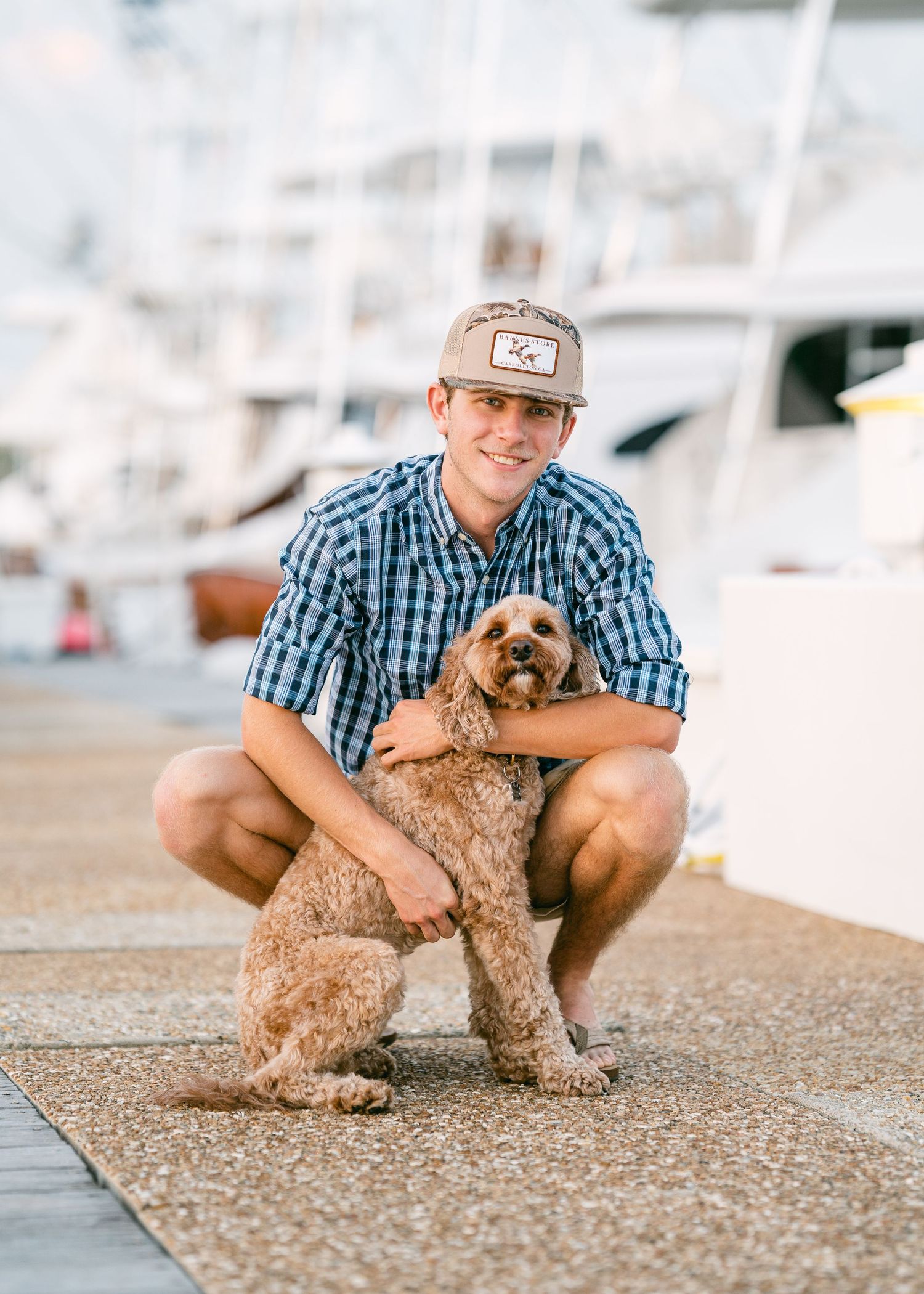 Smiling young man in plaid shirt crouches with fluffy brown dog at marina dock.