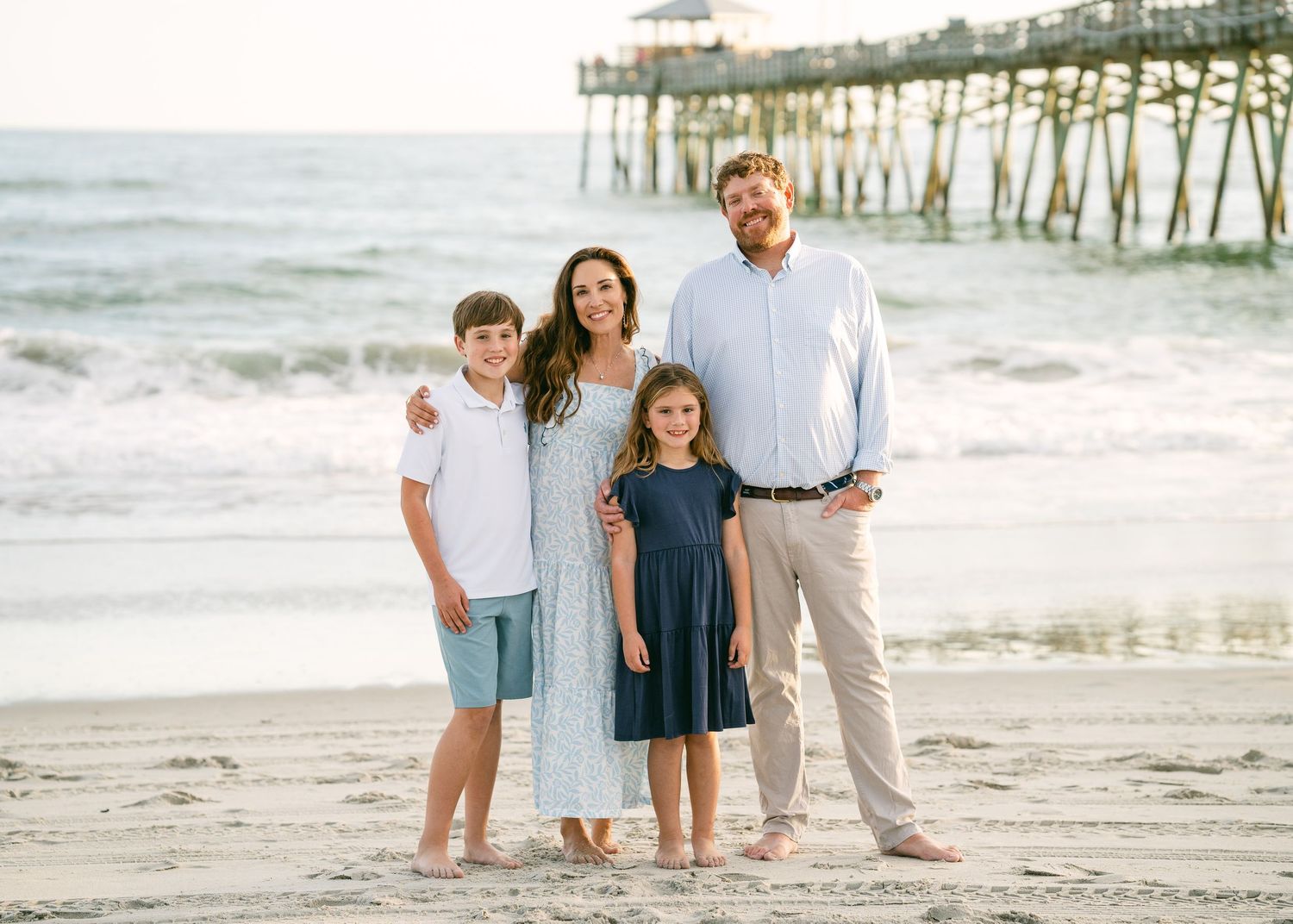 Happy family of four posing together on sandy beach near pier.