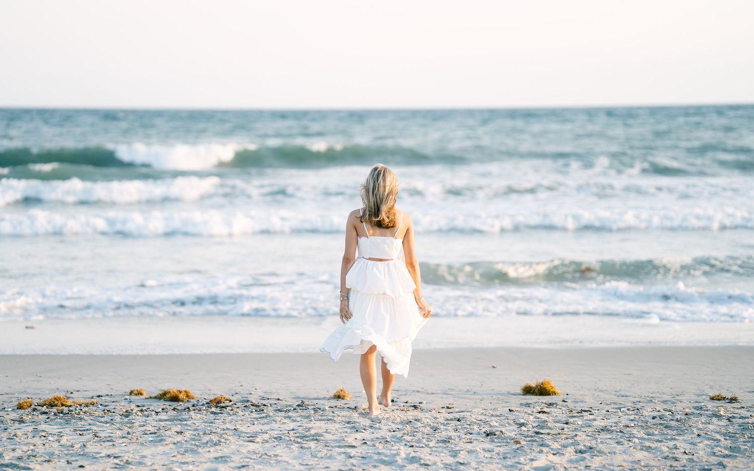 Woman in white dress walking toward ocean waves on sandy beach from behind.