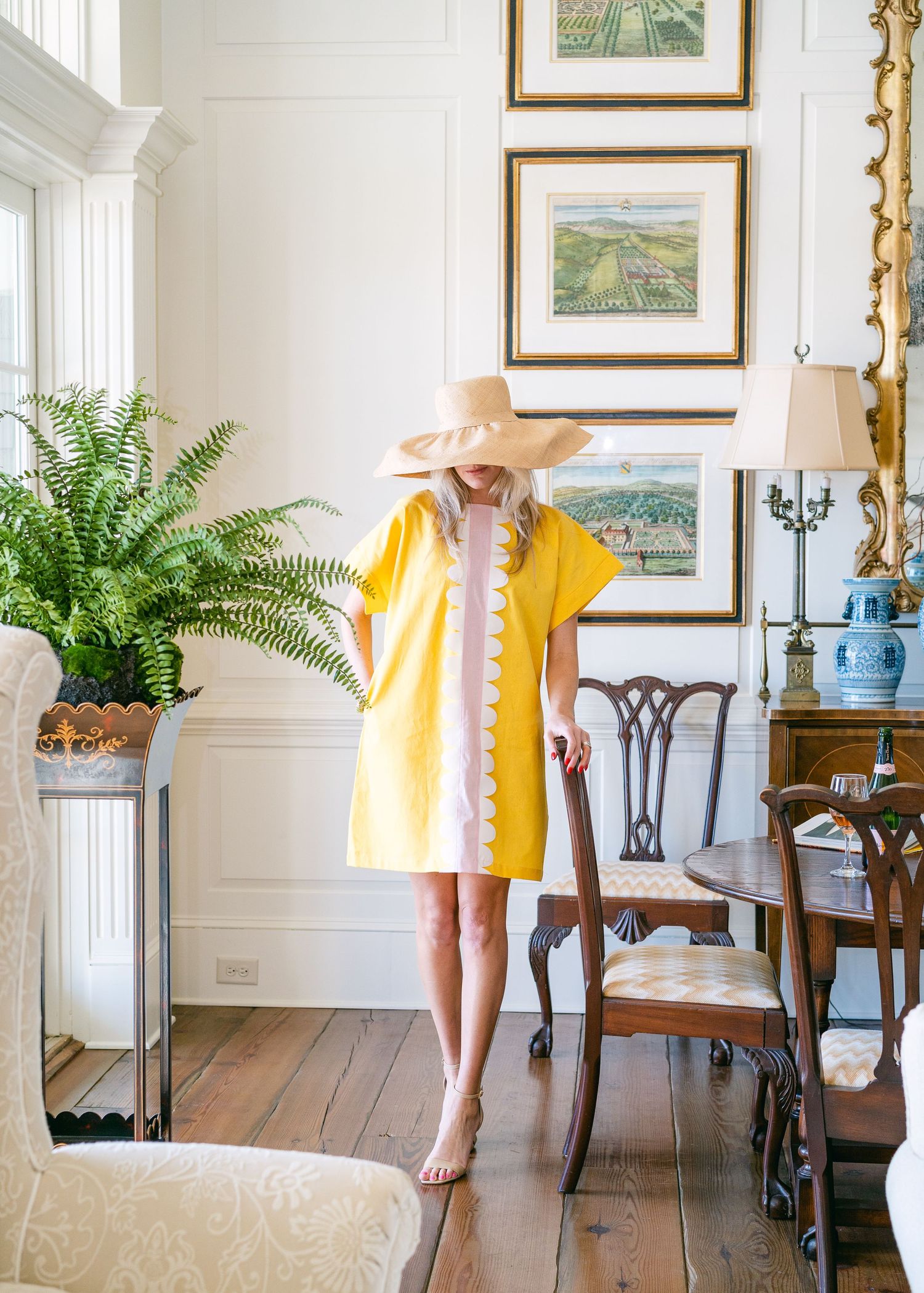 Woman in yellow shift dress and wide-brim hat standing in bright elegant dining room.