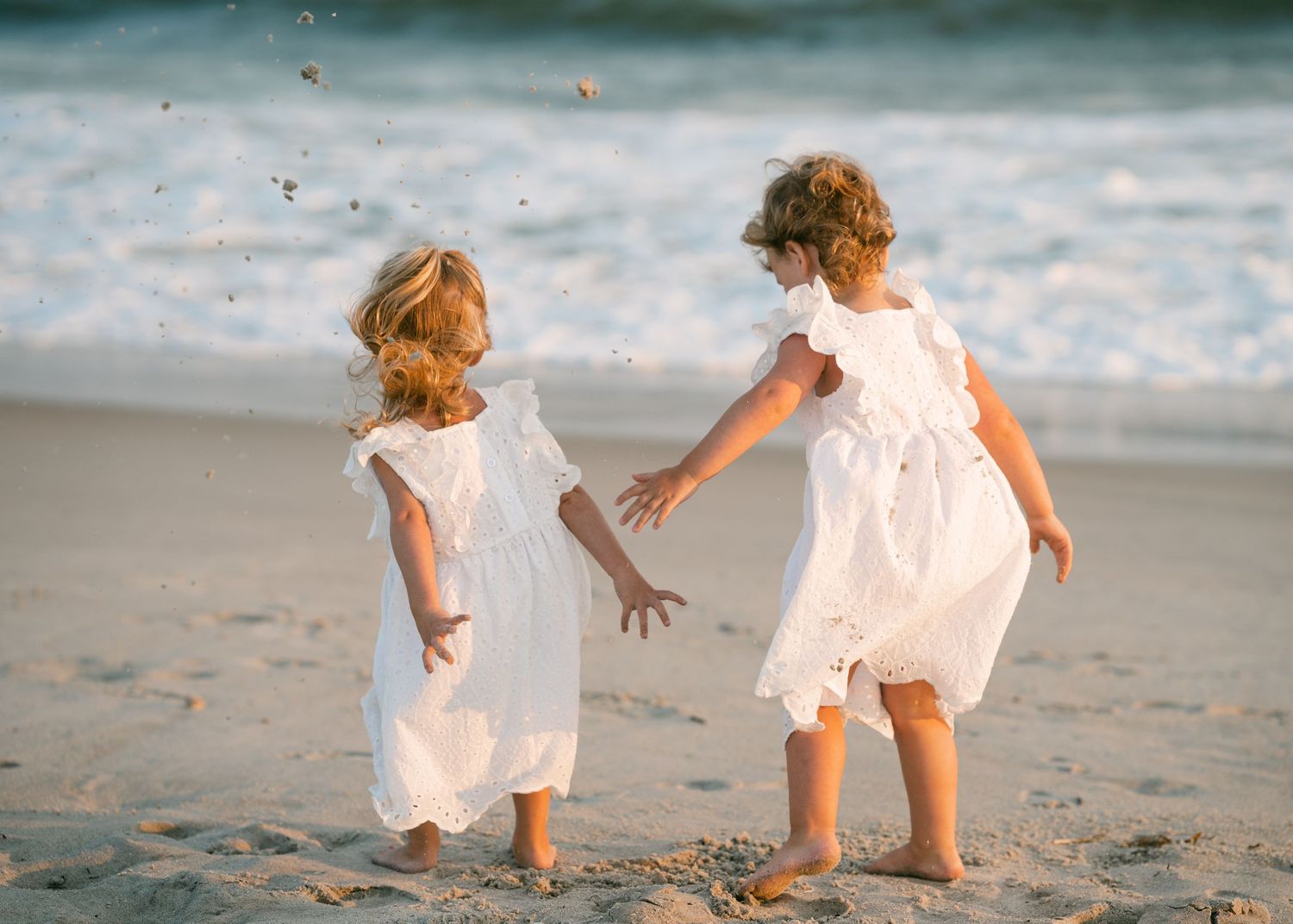 Two little girls in white dresses playing on a sunny beach together.