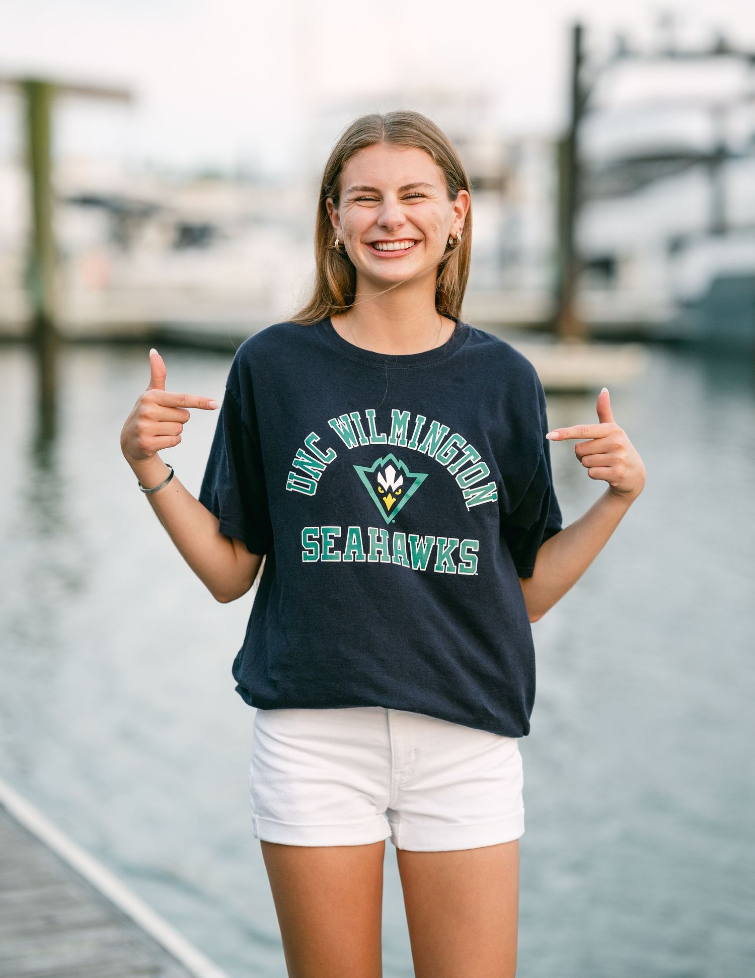 Laughing young woman pointing to UNC Wilmington Seahawks navy shirt at marina waterfront.