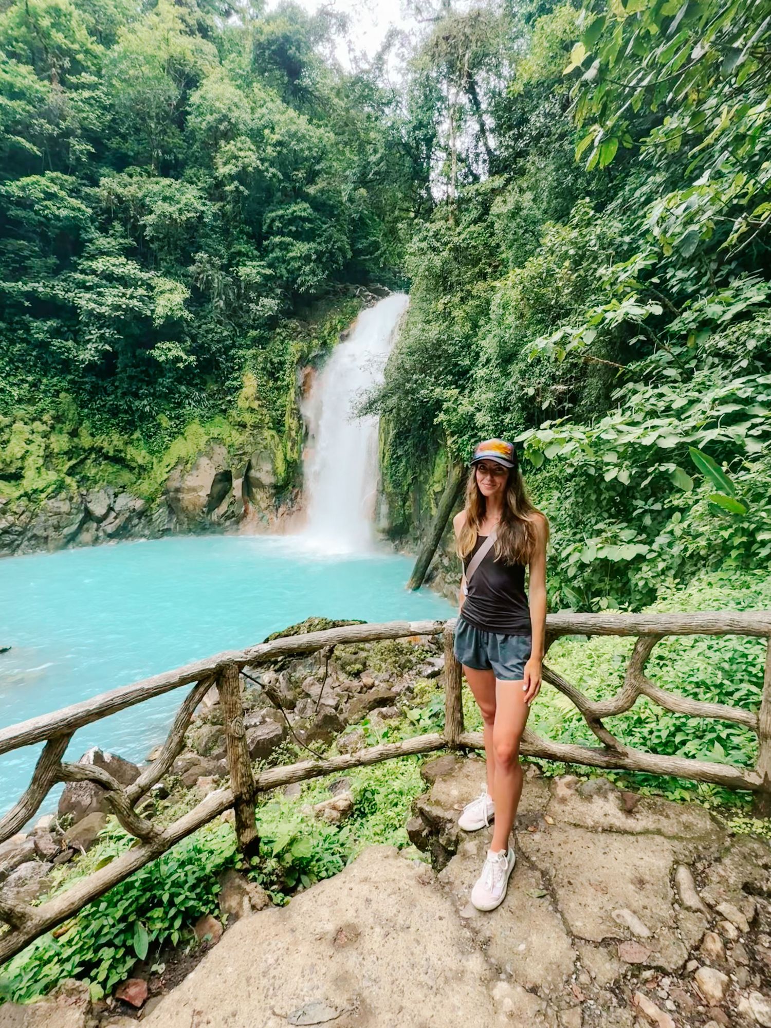 Woman poses at wooden railing overlooking stunning turquoise waterfall in Costa Rican rainforest.