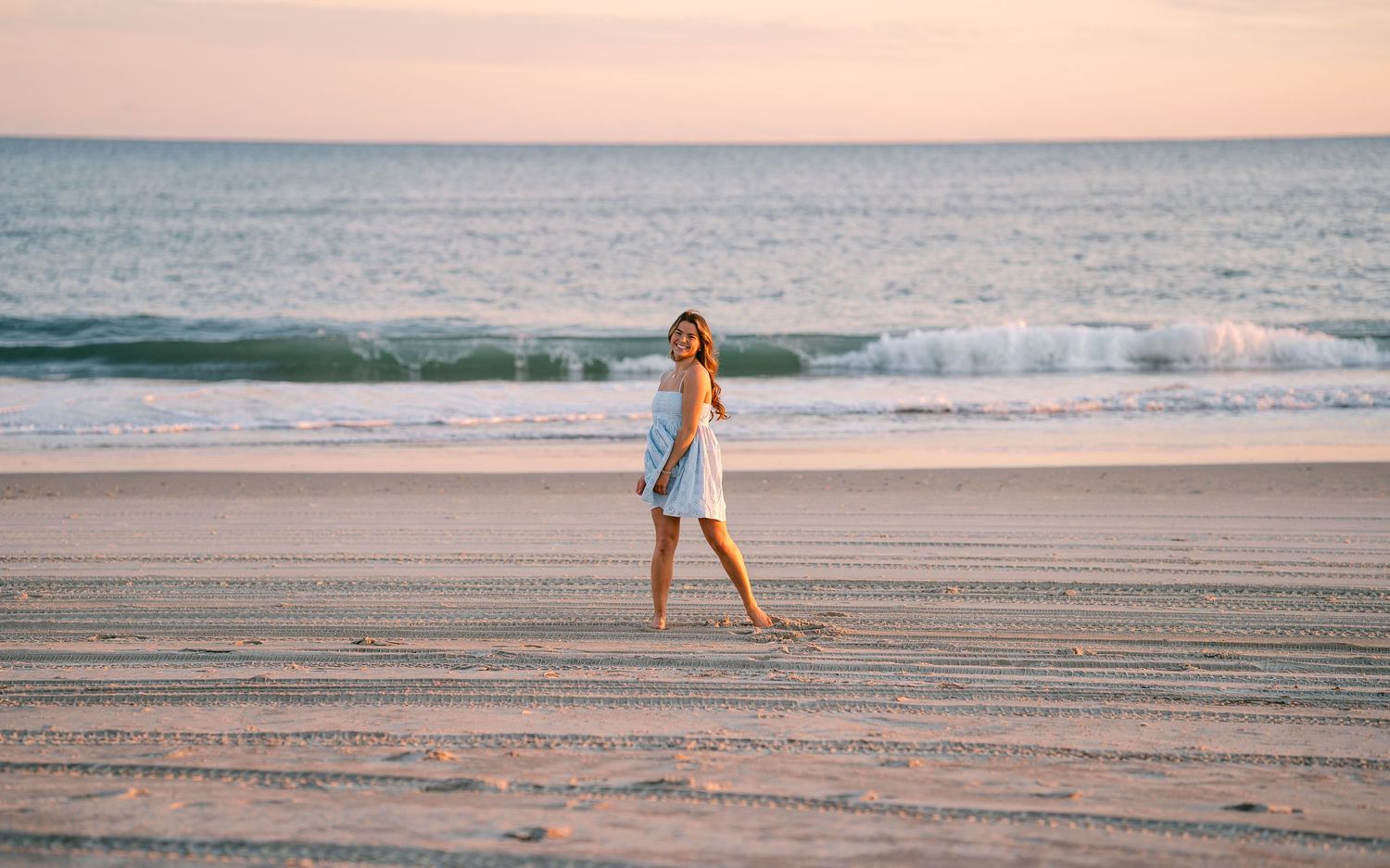 Young woman in white dress walking alone on a wide sandy beach at pastel sunset.