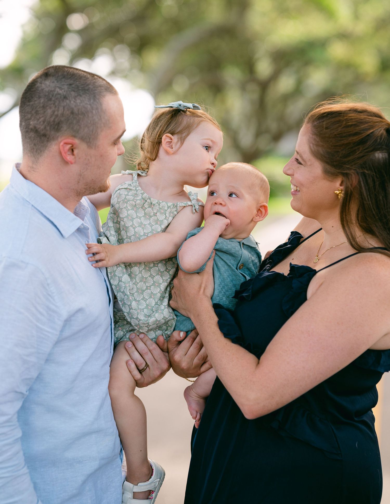 Parents lovingly watching toddler girl kiss baby brother outdoors under green trees.