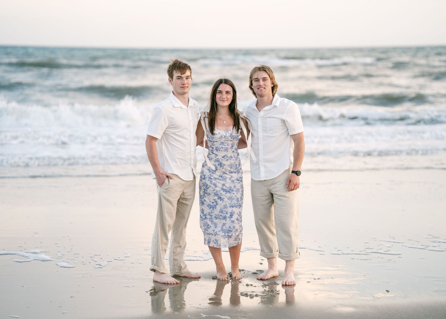 Three young adult siblings smiling together on beach with ocean waves behind them.