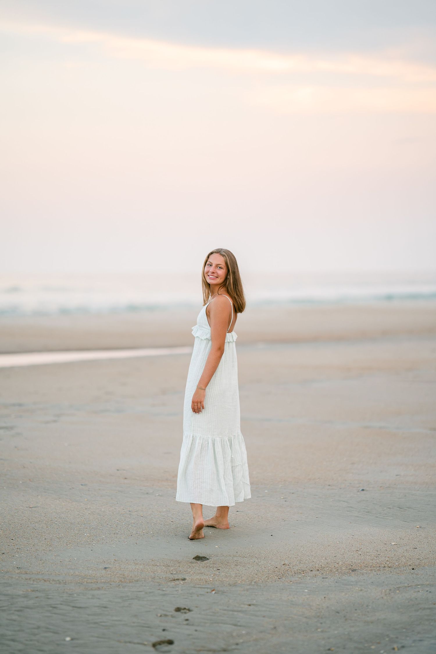 Young woman in white maxi dress walking barefoot on sandy beach at soft pastel sunset.