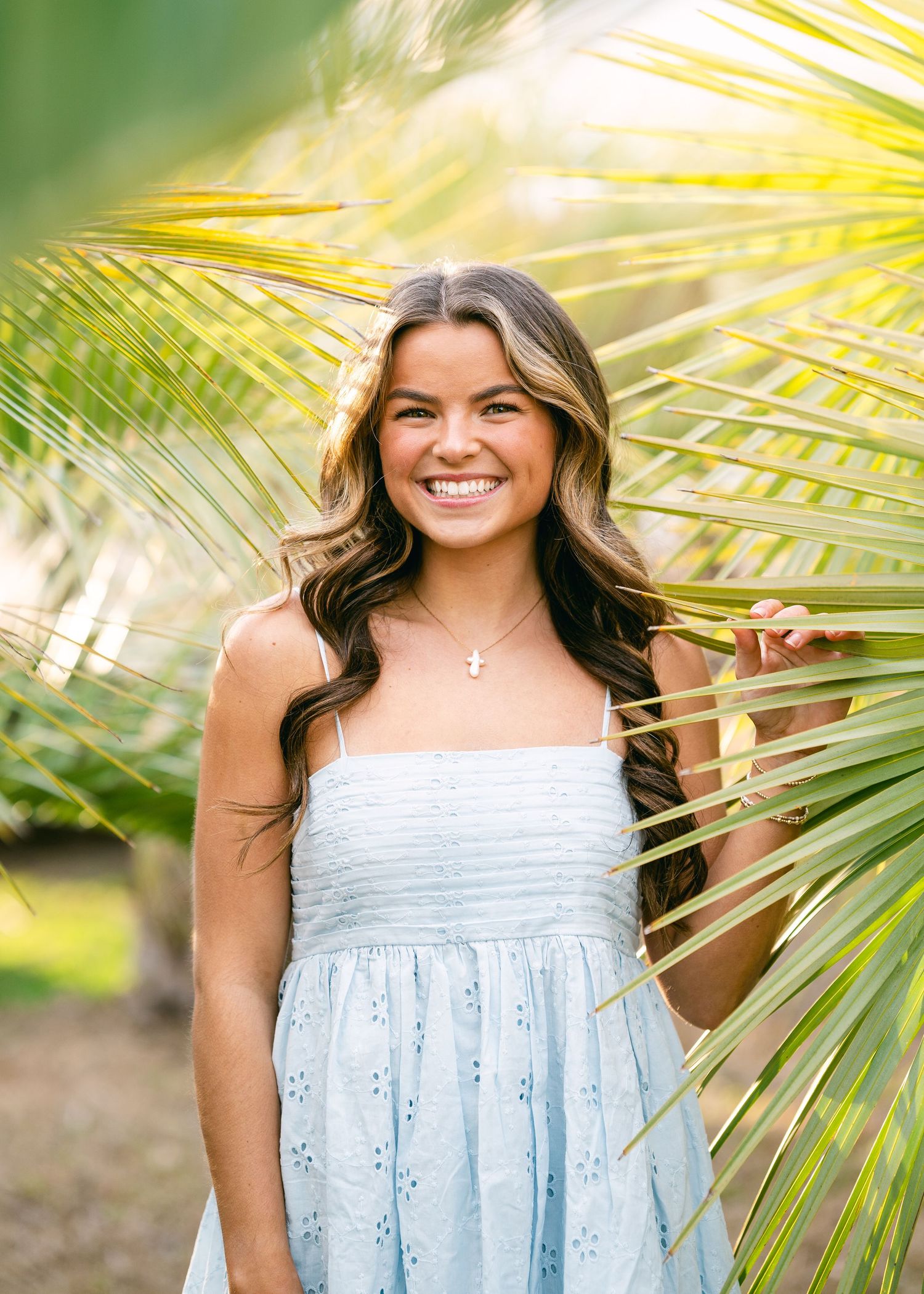 Smiling teenage girl in light blue sundress standing among green palm fronds in sunny outdoor setting.