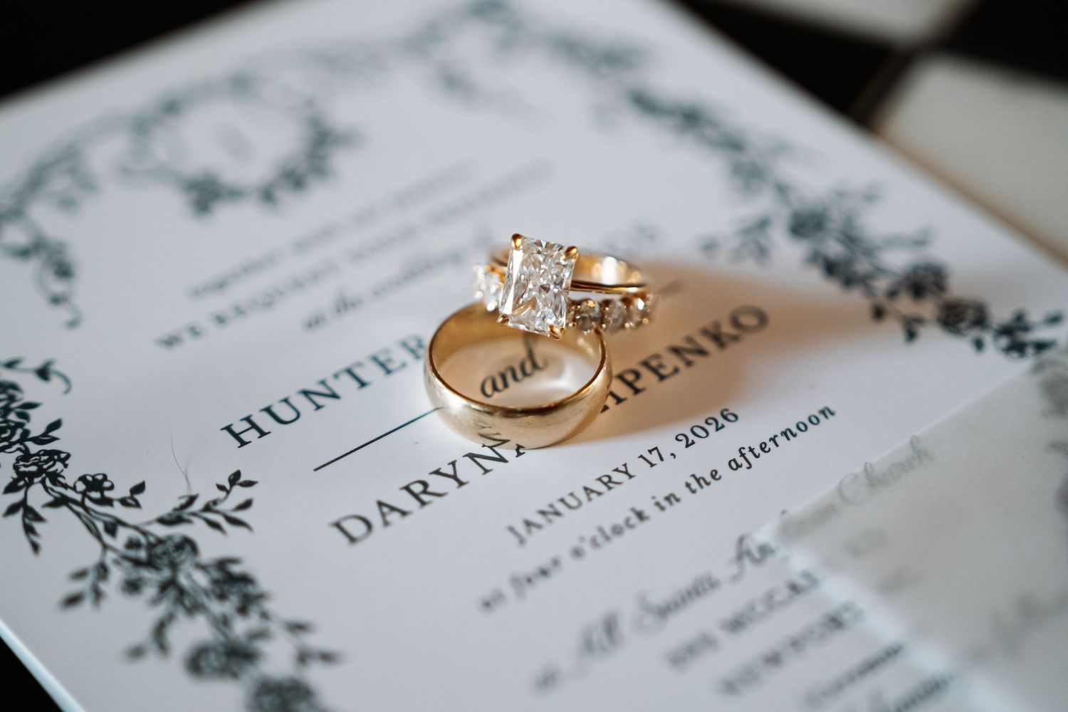 Gold wedding bands and diamond engagement ring displayed on elegant floral wedding invitation dated January 17 2026.