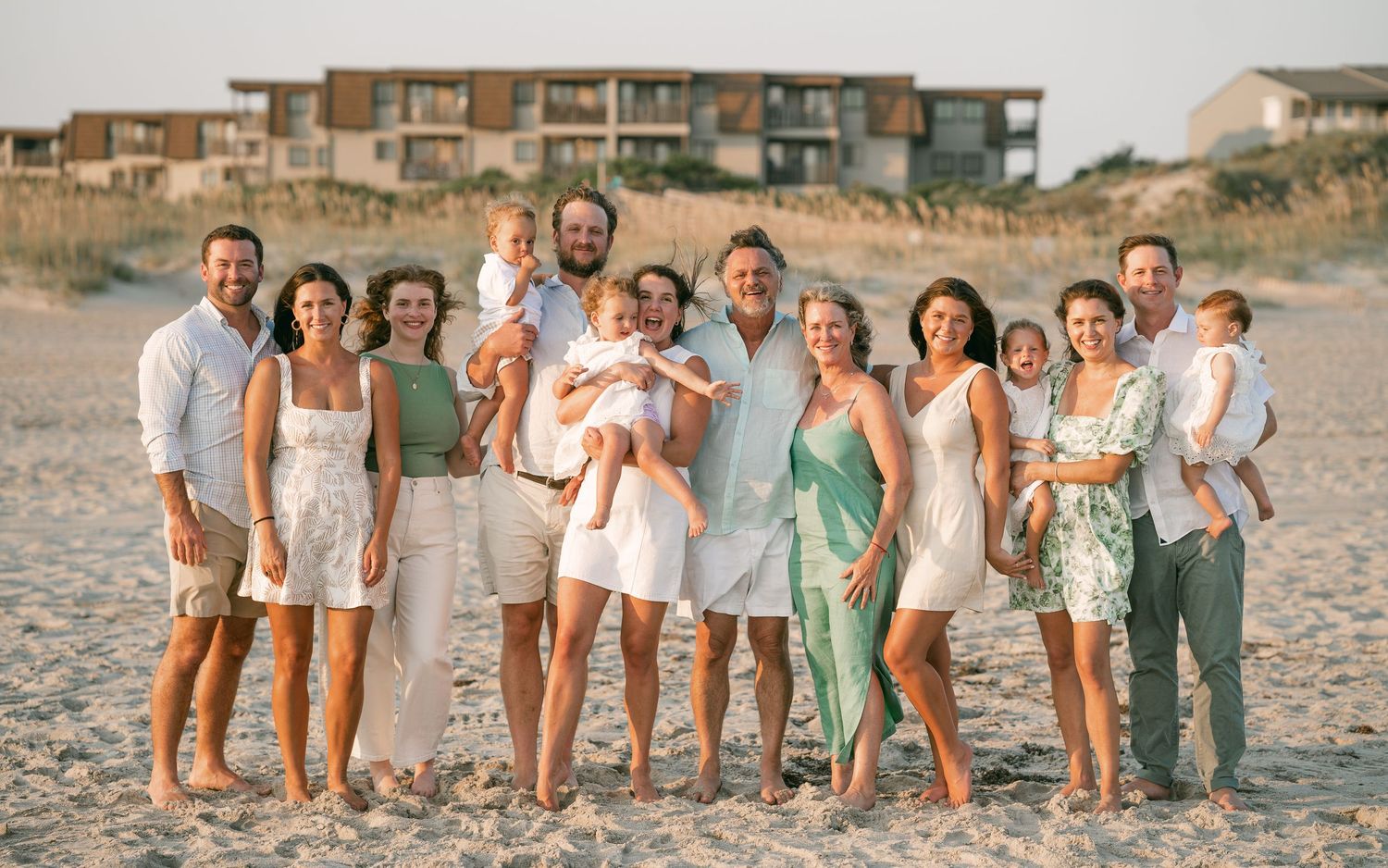 Large family group photo on beach, adults and children in coordinating white and green outfits, smiling together near ocean.