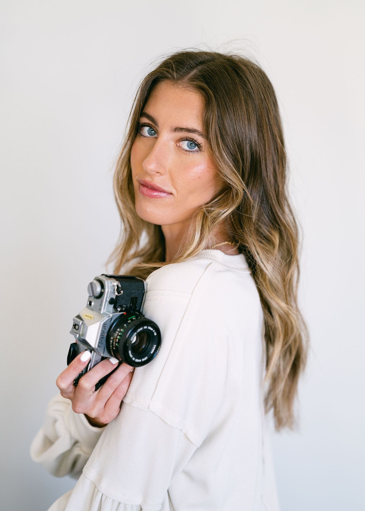Young woman with long wavy brown hair holds a vintage film camera, wearing a cream top against a white background.