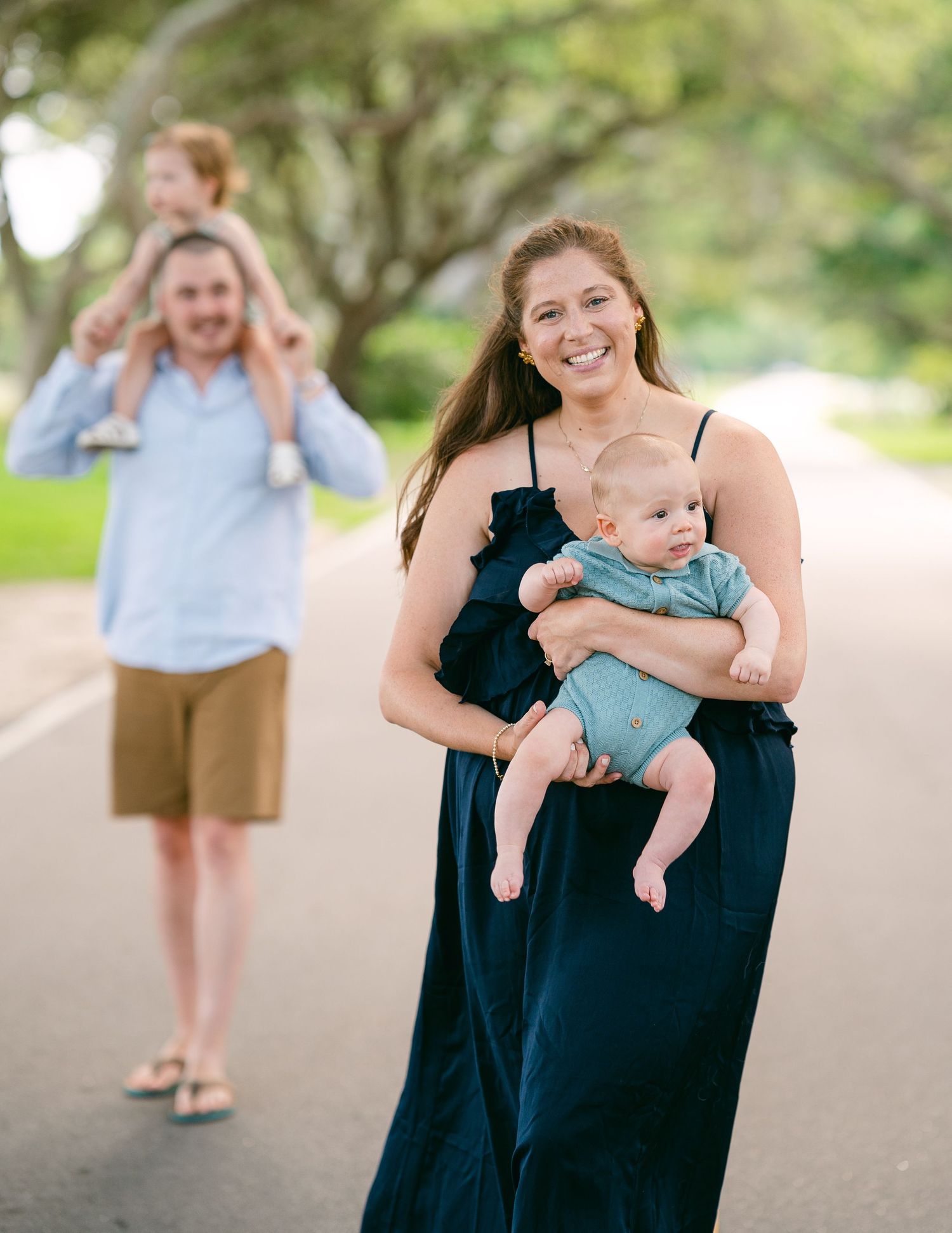 Smiling mother holding infant on tree-lined road while father carries toddler on shoulders.