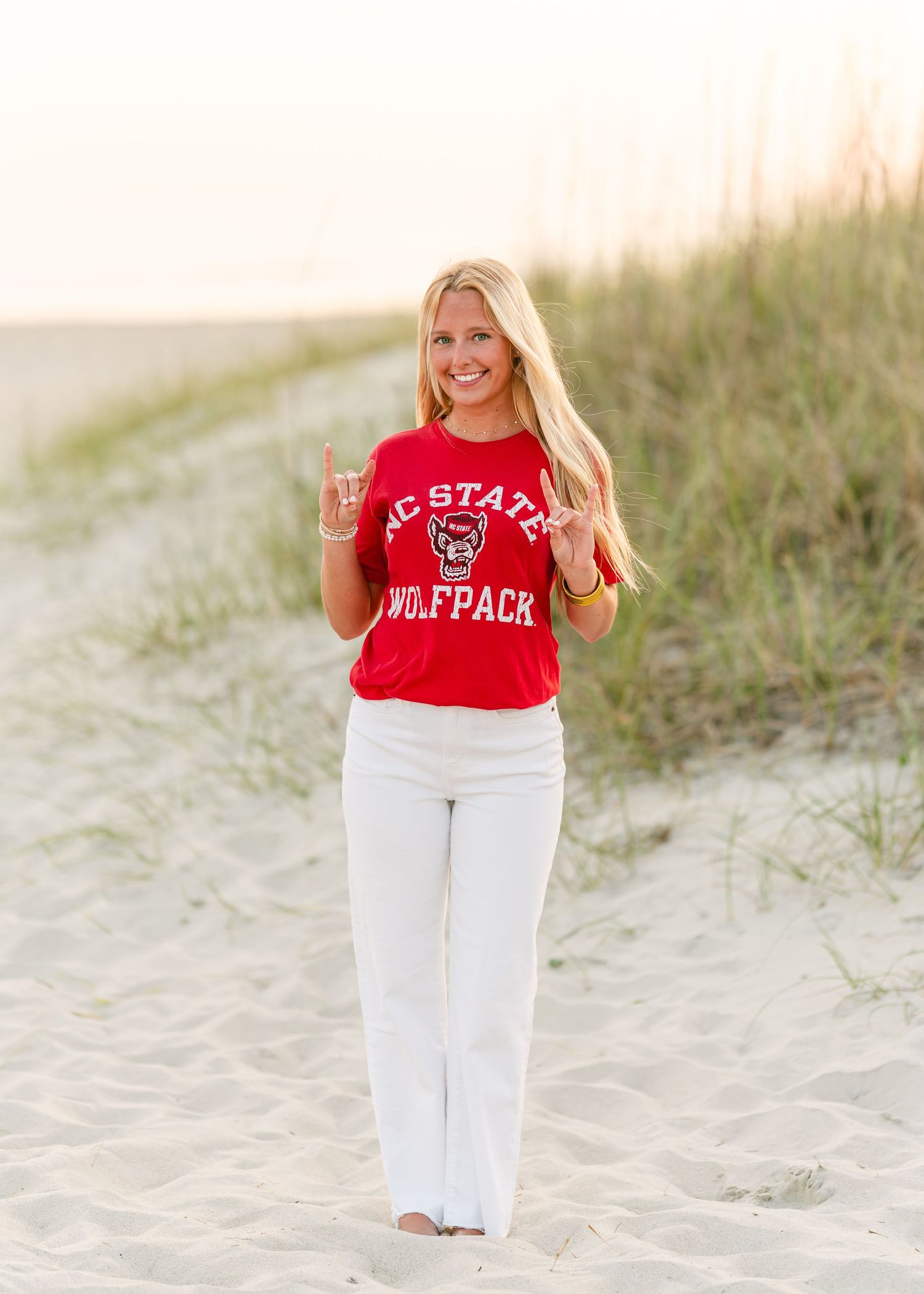 Smiling blonde woman in NC State Wolfpack red shirt posing on sandy beach at golden hour.