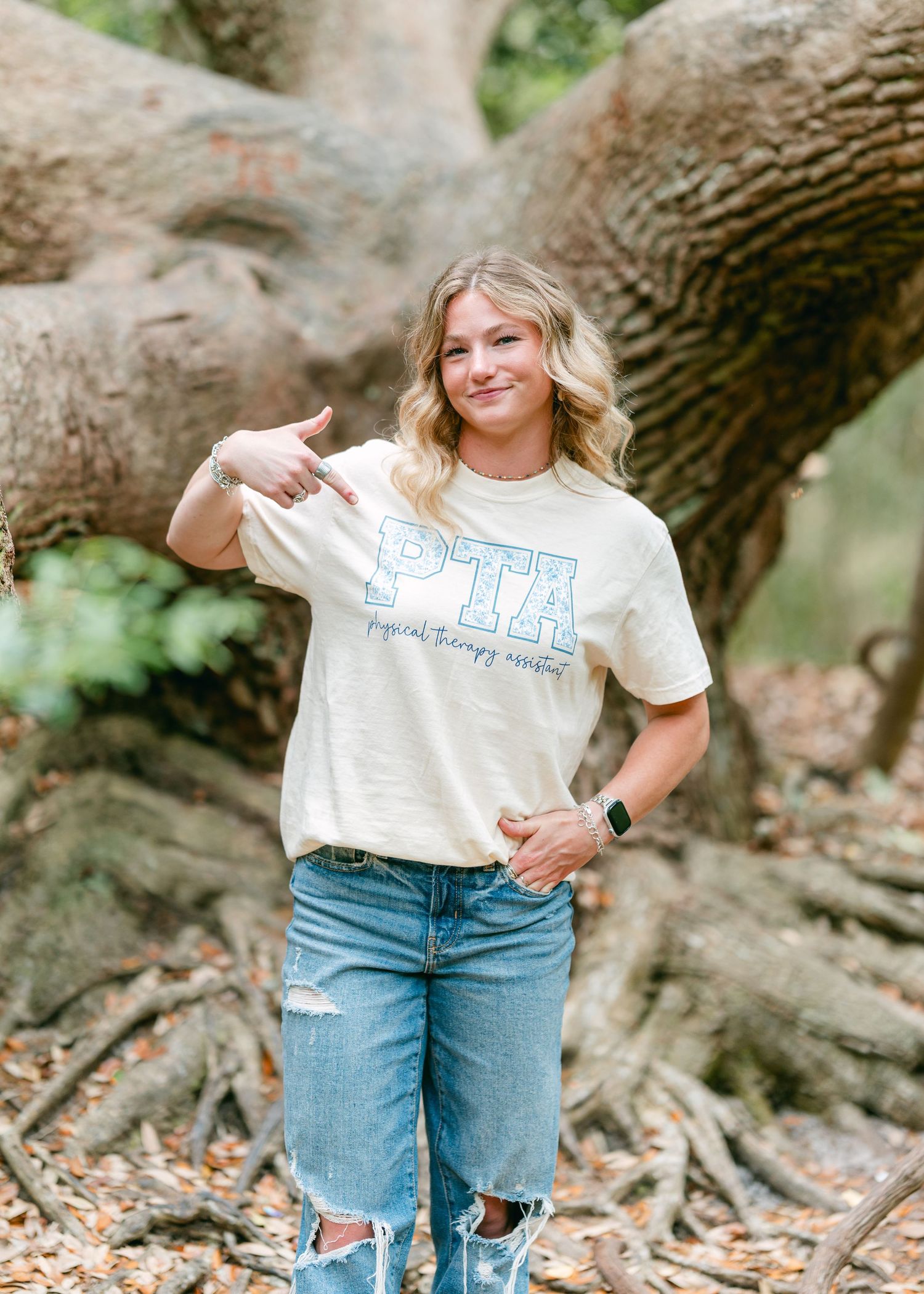 Smiling blonde woman pointing to PTR physical therapy assistant t-shirt near large tree roots.