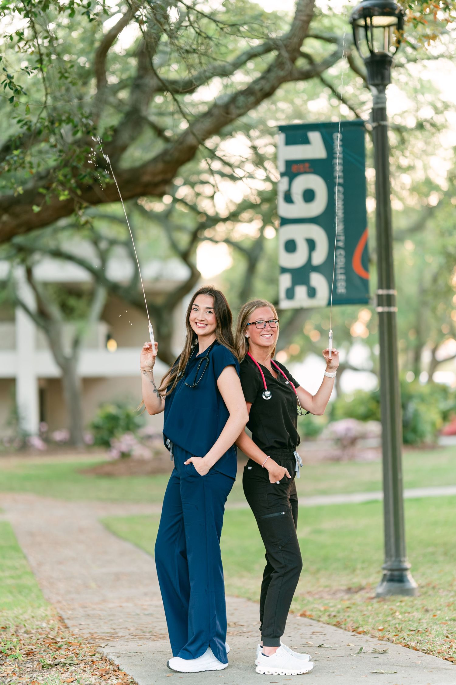 Two nursing students in scrubs pose back-to-back near a 1963 university banner outdoors.
