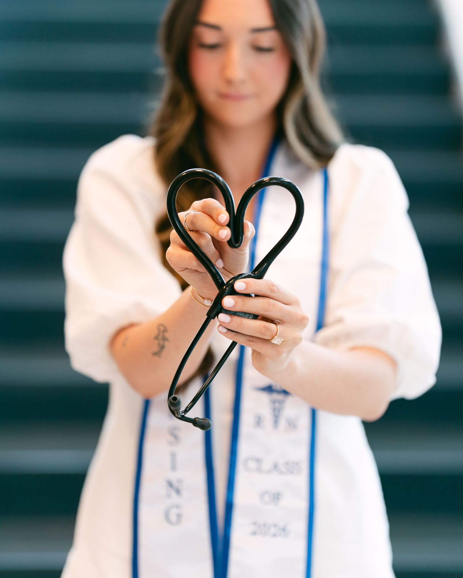 Nursing graduate in white coat holding stethoscope in heart shape for Class of 2026 photo.