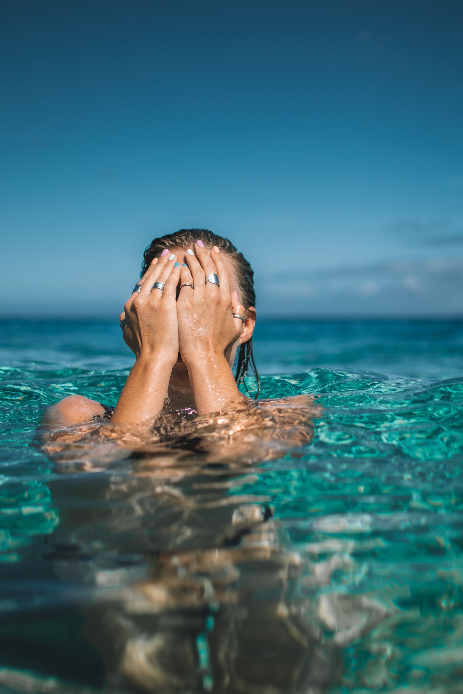 Woman covers face with hands while emerging from crystal clear turquoise tropical ocean water.