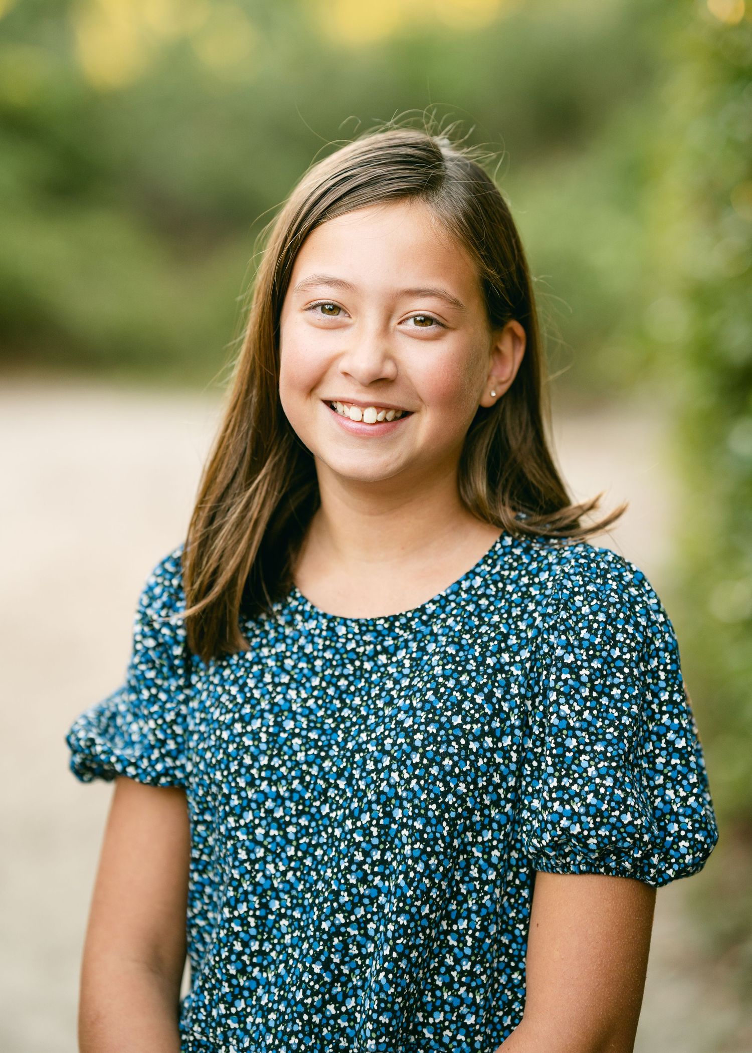 Smiling preteen girl with brown hair wearing a blue floral dress outdoors.