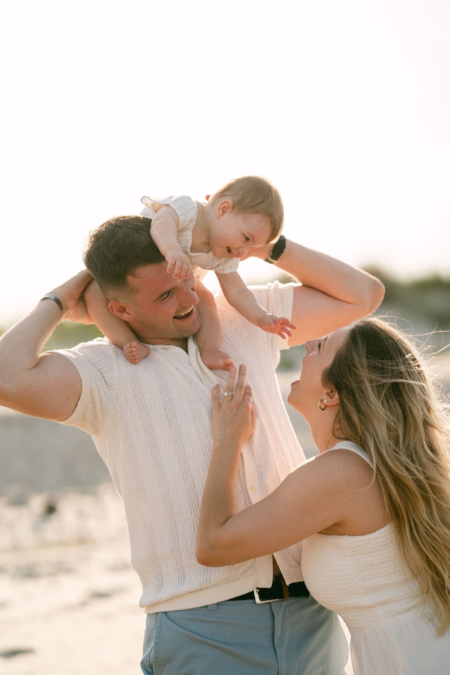 Joyful parents lifting giggling baby above their heads on bright sunny beach.