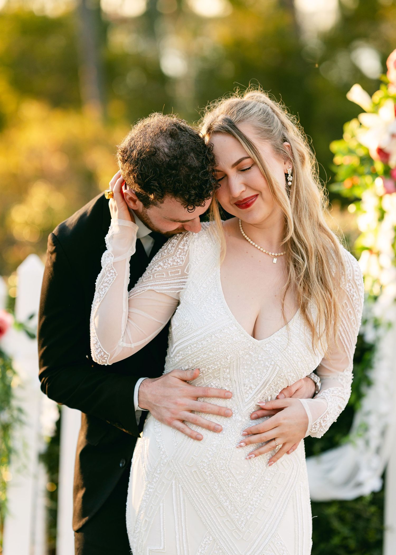 Groom embraces pregnant bride in beaded gown during outdoor wedding ceremony at golden hour.