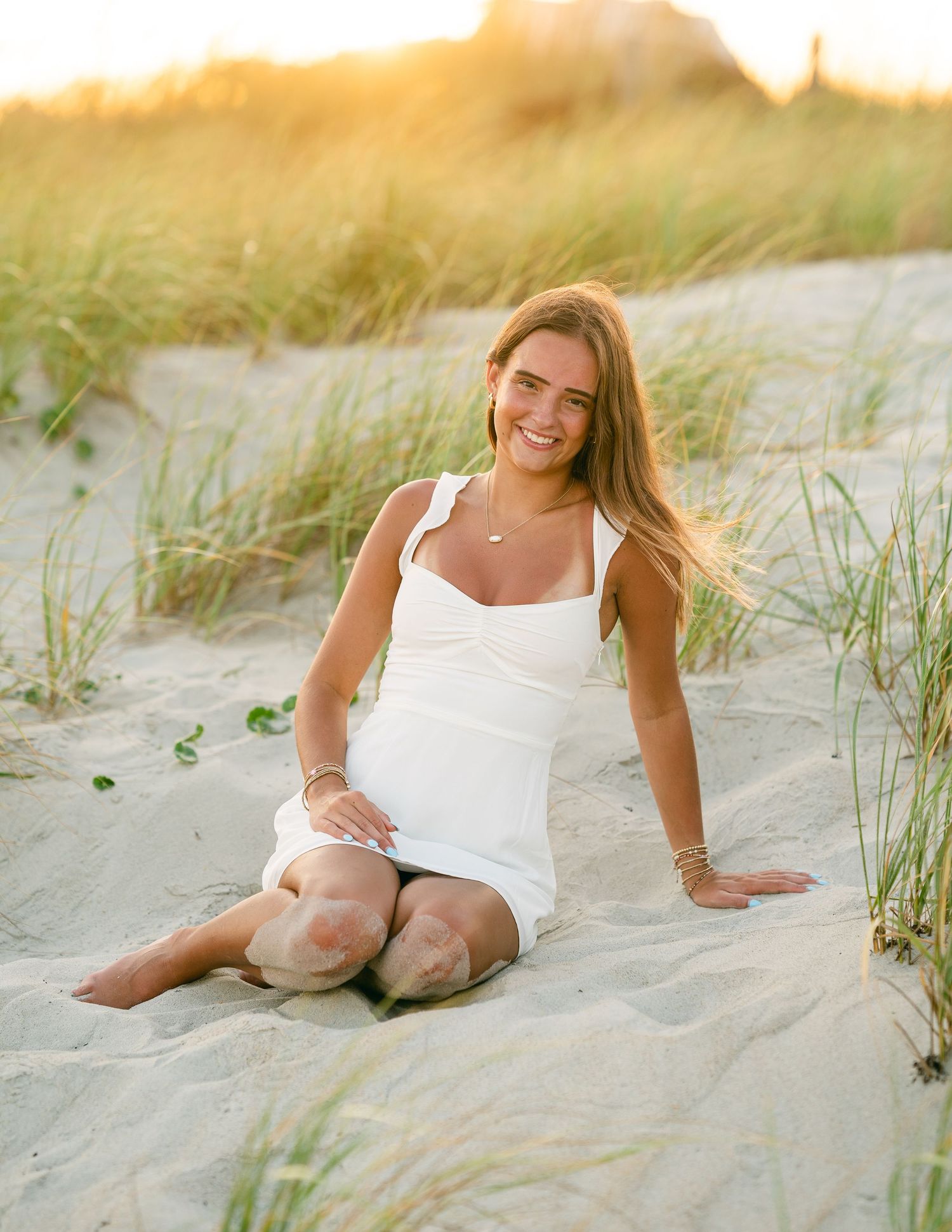 Smiling young woman in white dress sitting on sandy beach dunes at golden sunset hour.