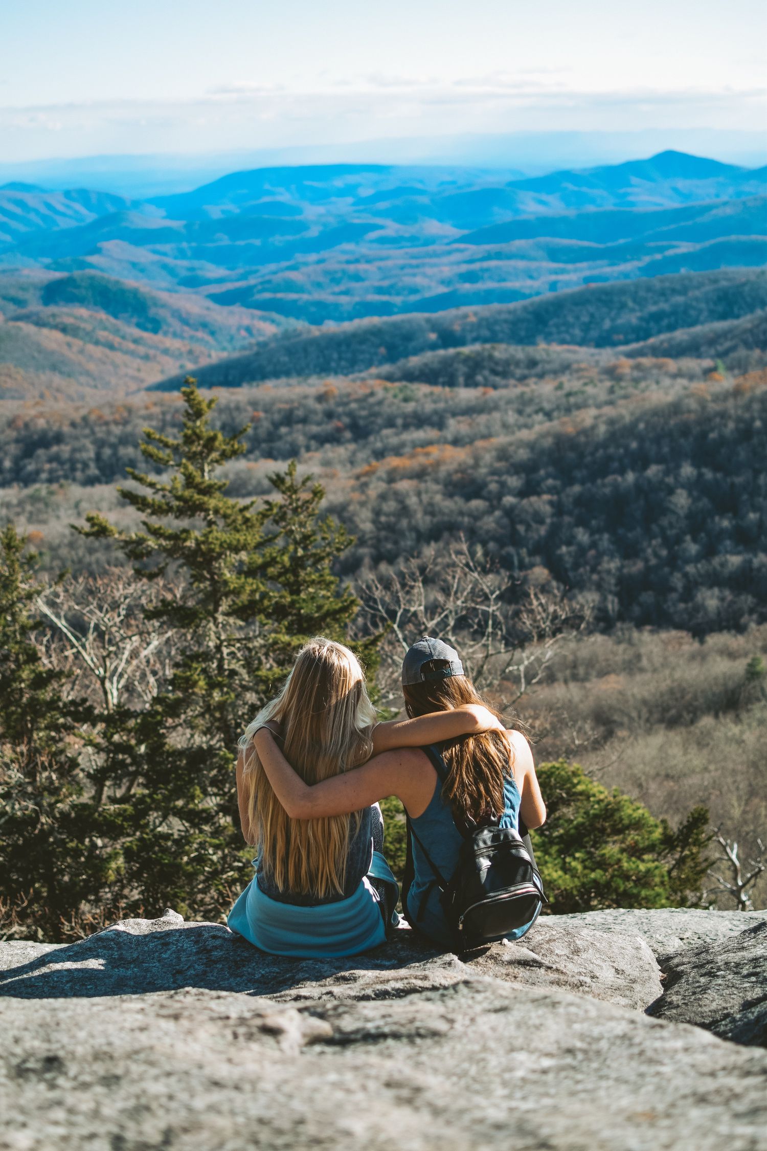Two friends sitting on a mountain rock ledge with arms around each other enjoying panoramic valley views.