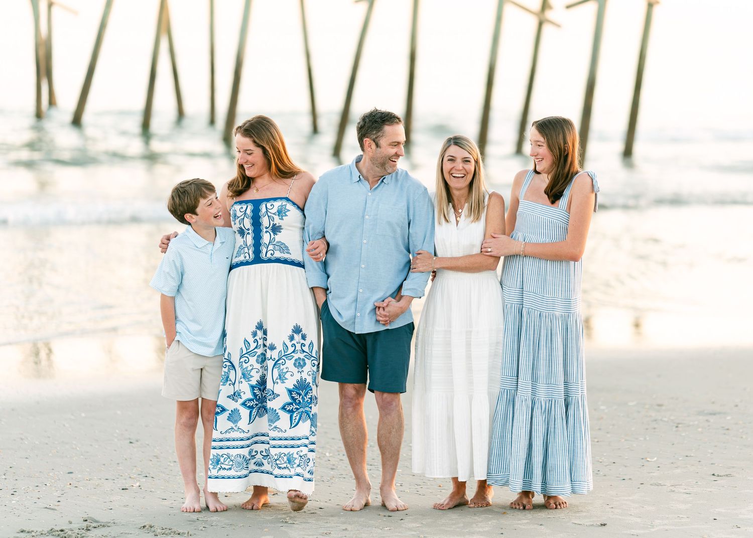 Cheerful family of five laughing together on beach near wooden pier at sunset.