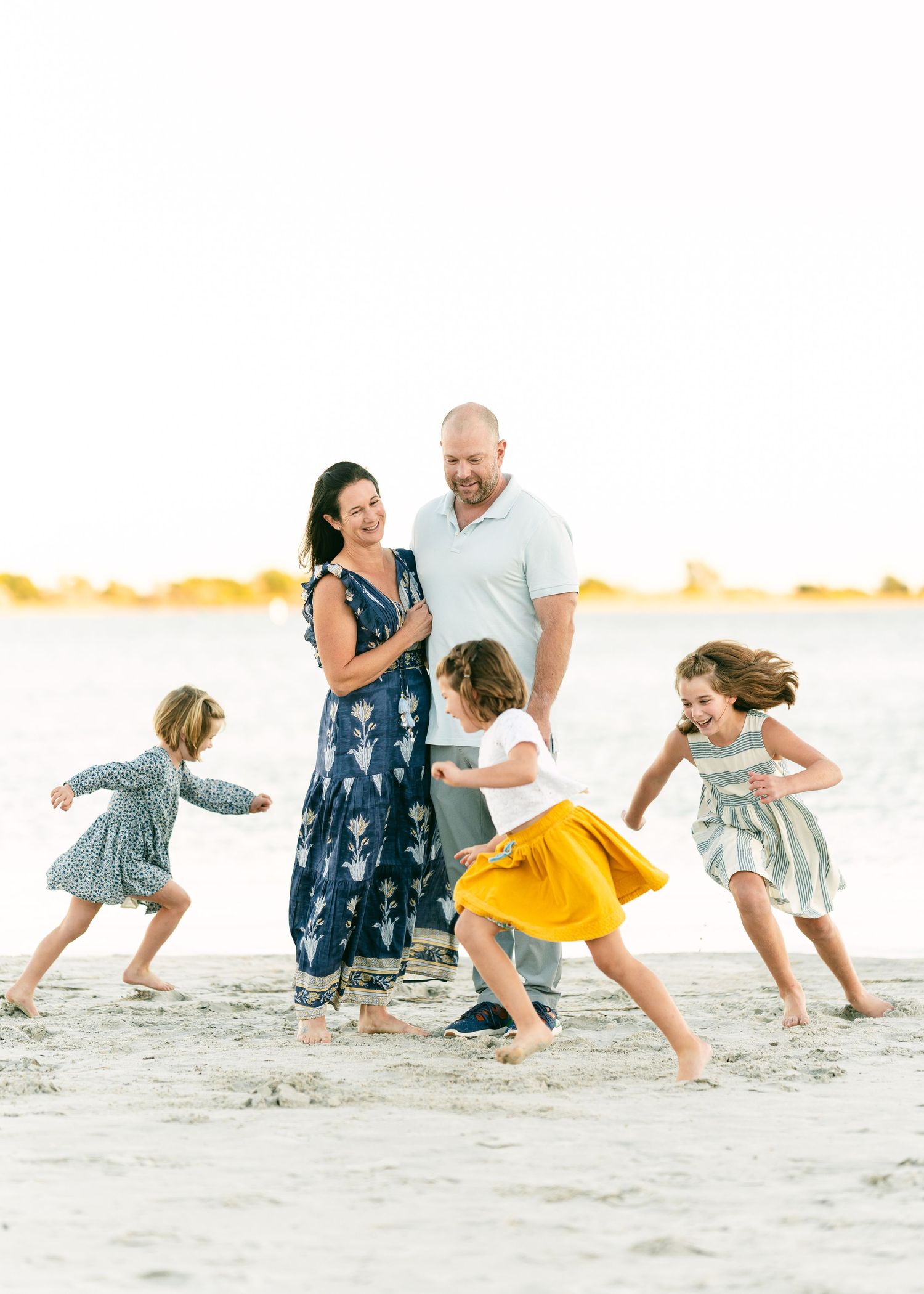 Parents watching three young daughters dancing playfully on bright sandy beach together.