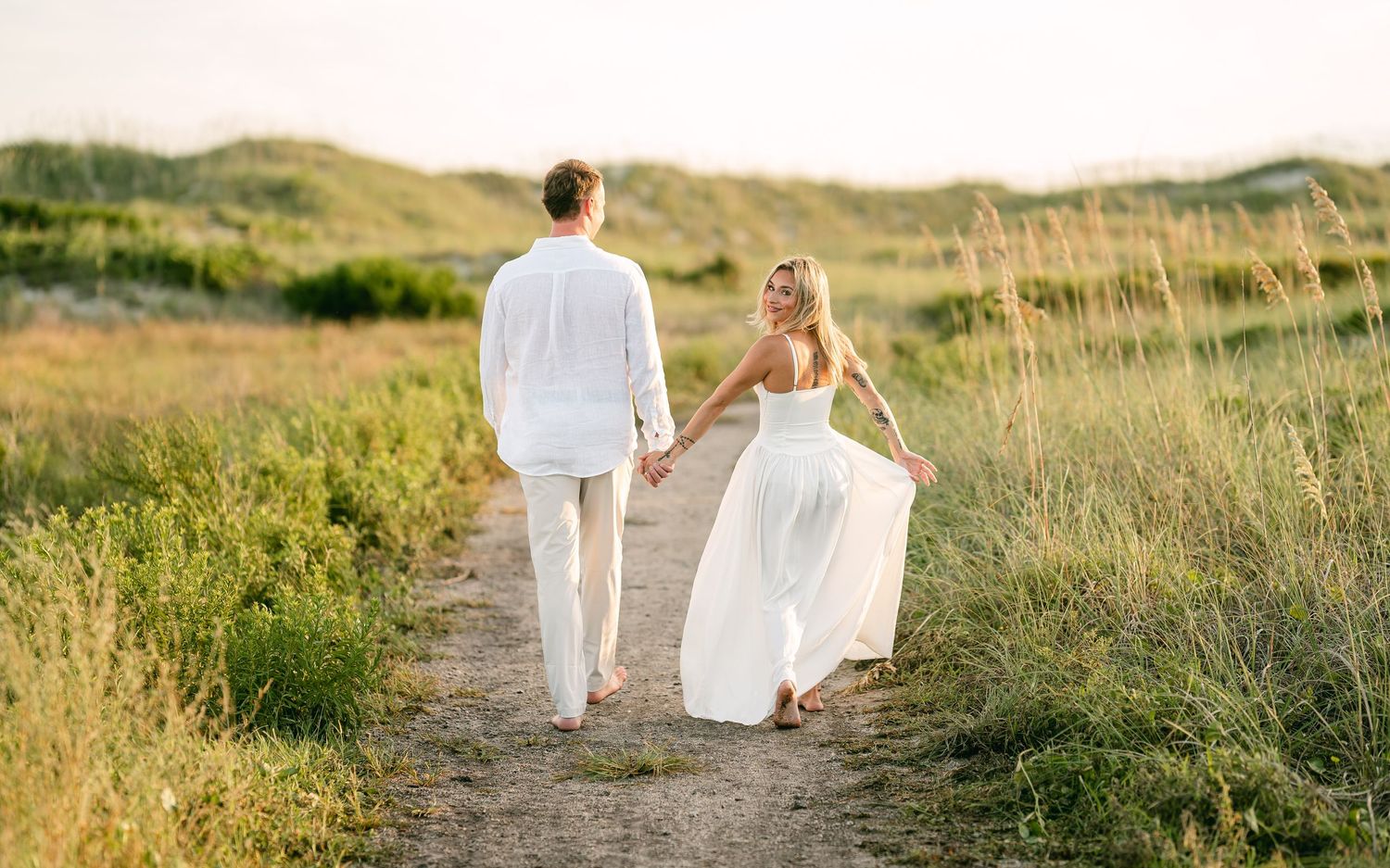 Couple in white outfits holding hands walking along a scenic coastal dune path at sunset.