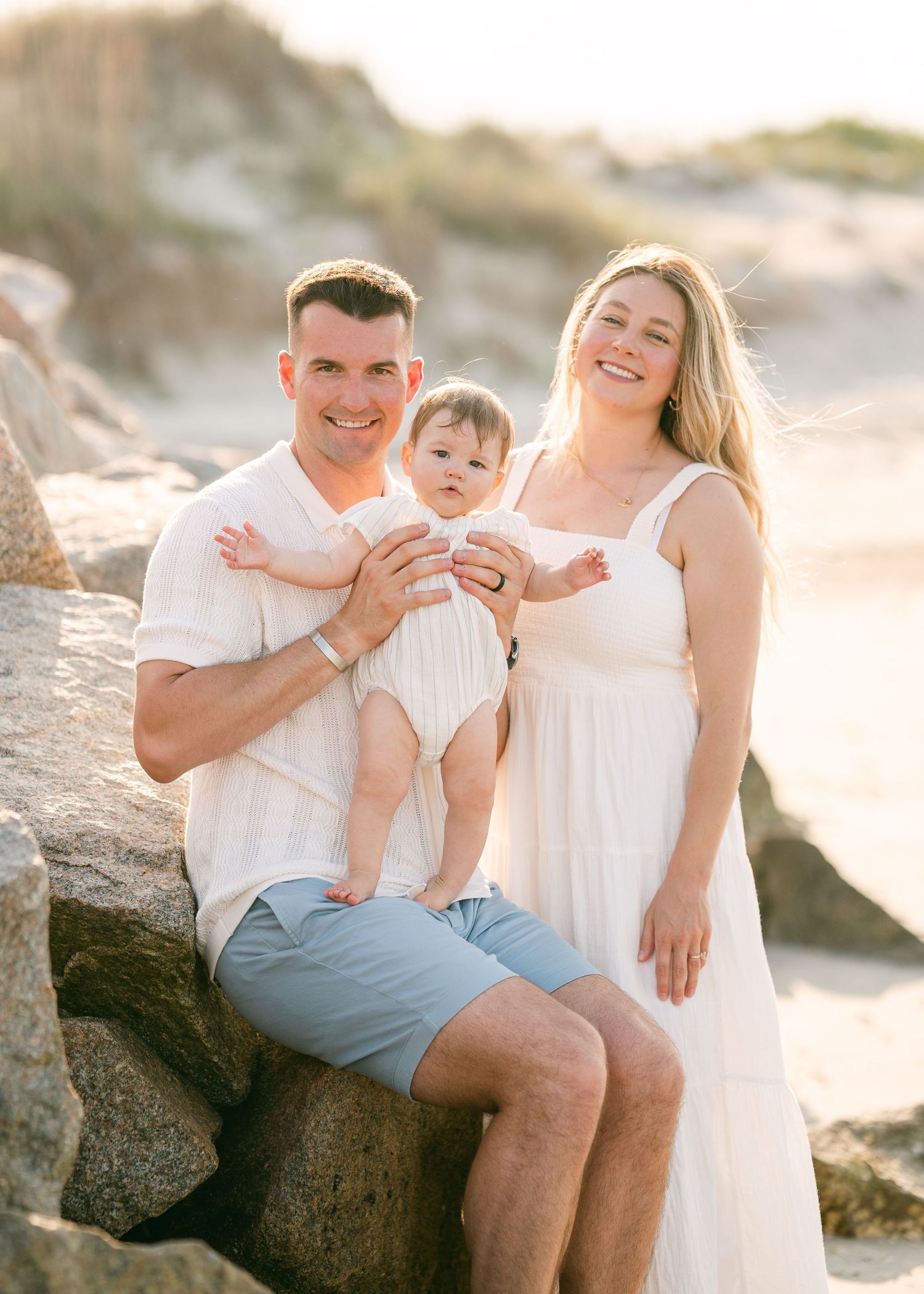Happy family sitting on beach rocks, father holding baby in white romper, mother in white dress.
