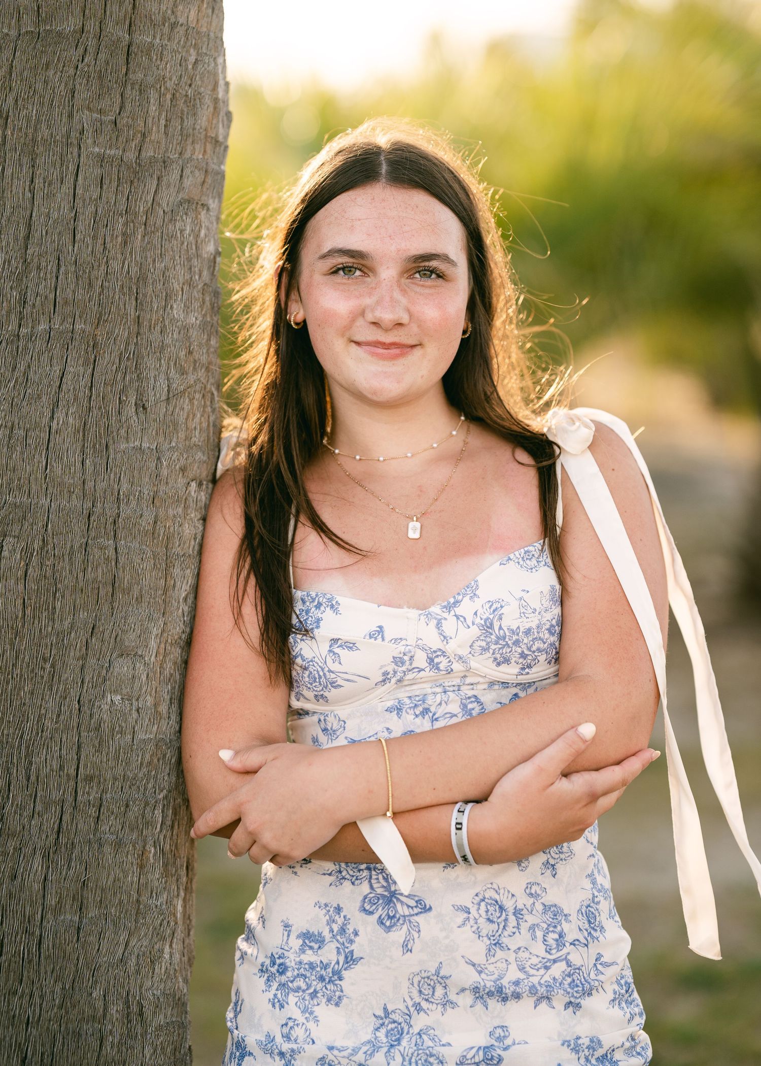 Smiling teenage girl in blue floral dress leaning against a palm tree in golden sunlight.