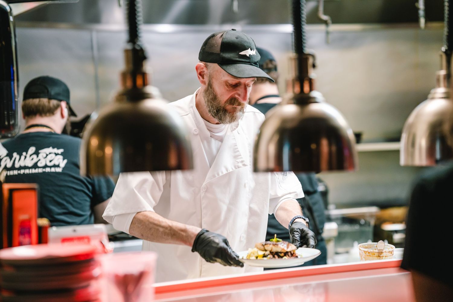 Chef in white coat plating food under heat lamps in a busy restaurant kitchen.