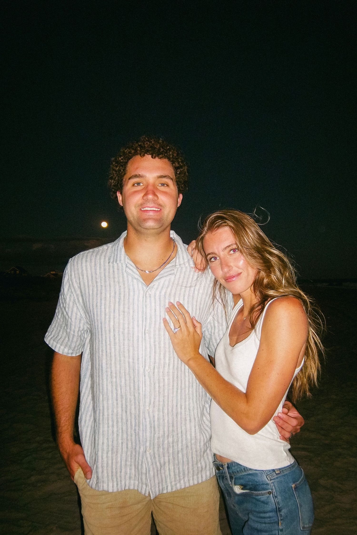 Couple embracing on a beach at night, woman showing engagement ring, full moon visible in sky.
